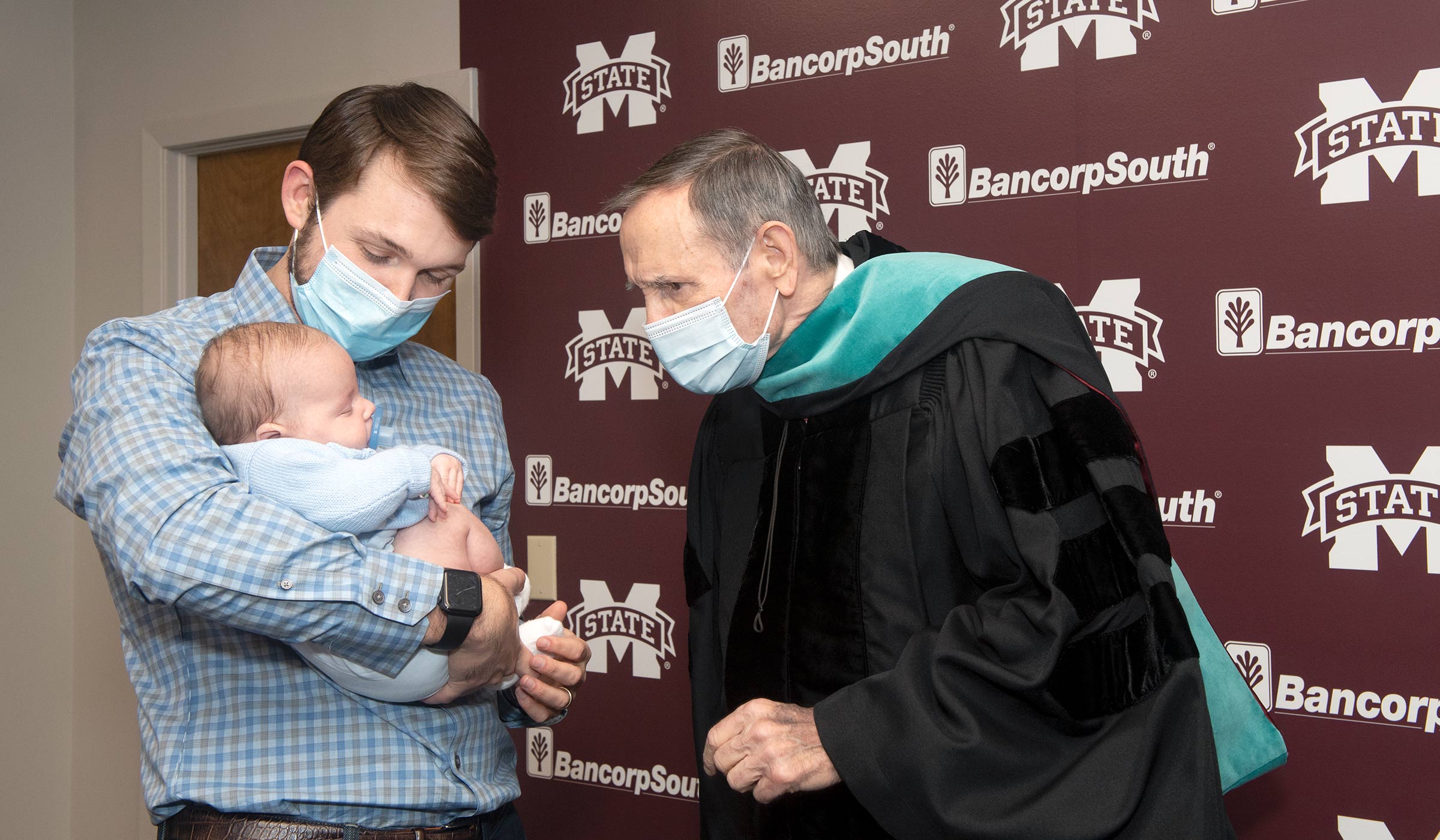 Man in mask and doctoral graduation gown looking at baby being held by man in checkered shirt.