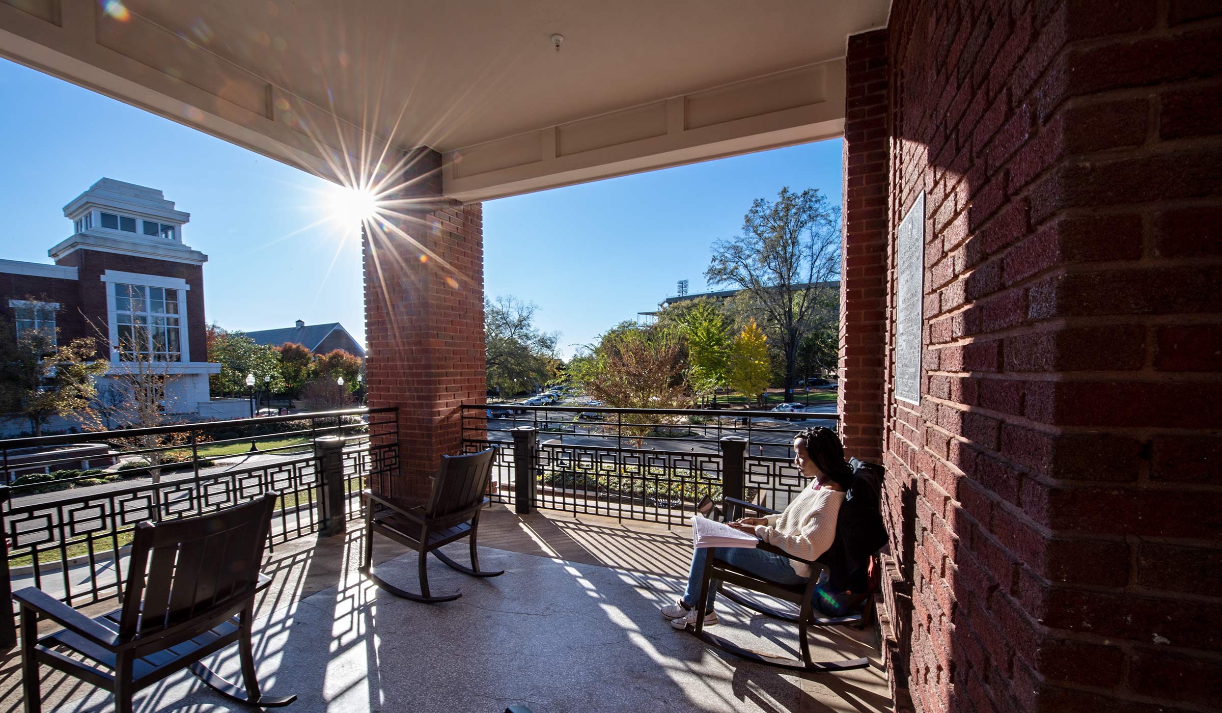 YMCA&#039;s front porch with rocking chairs, framing a view towards Colvard Tower. Student studies in a rocker in the sunshine.