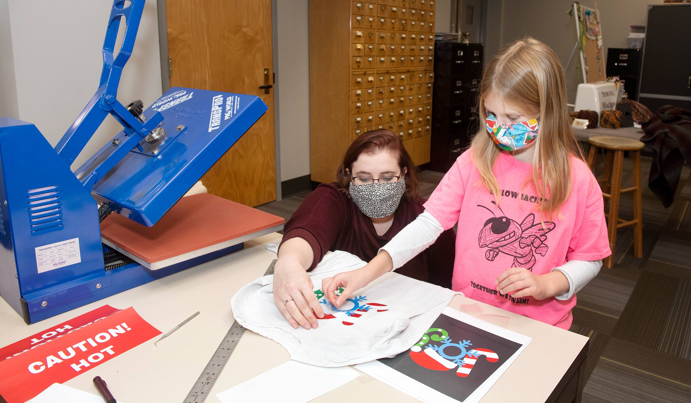 Girl in pink shirt and adult female in maroon peeling film off t-shirt near heat press