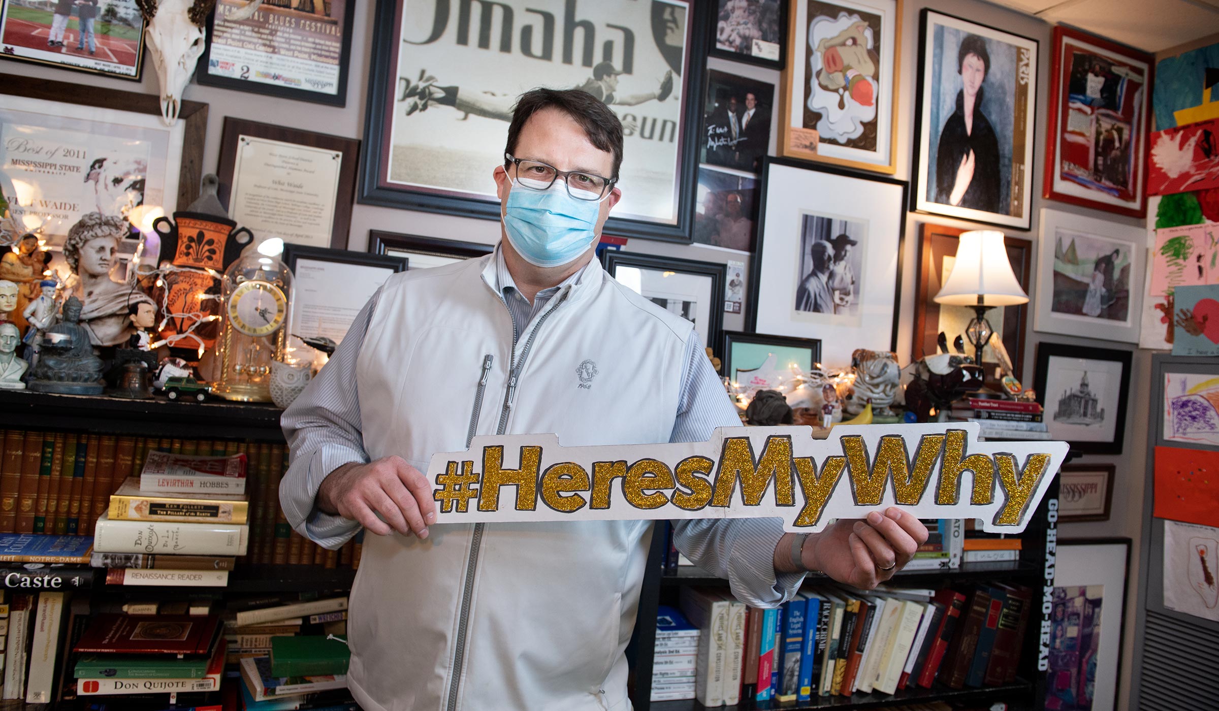 Poli Sci faculty member Whit Waide stands in his picture-covered office, wearing a facemask and holding a #HeresMyWhy sign.