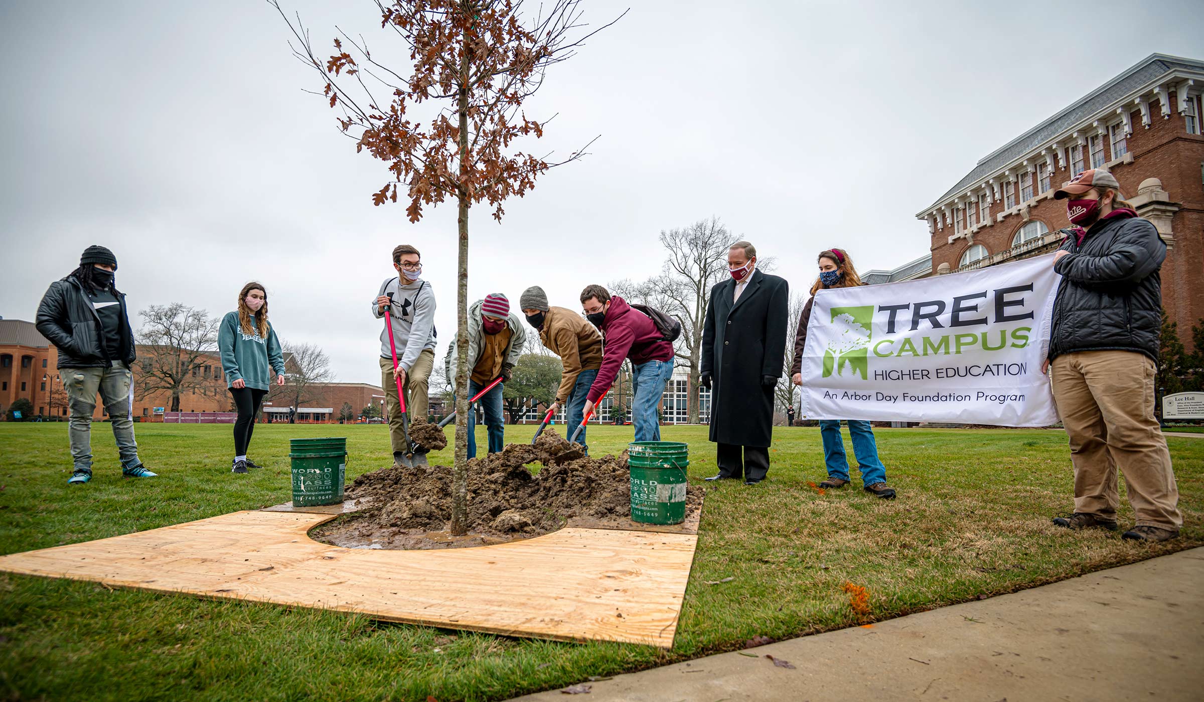 Students plant a tree on the Drill Field, with President Keenum looking on, along with two students holding a Tree Campus sign.