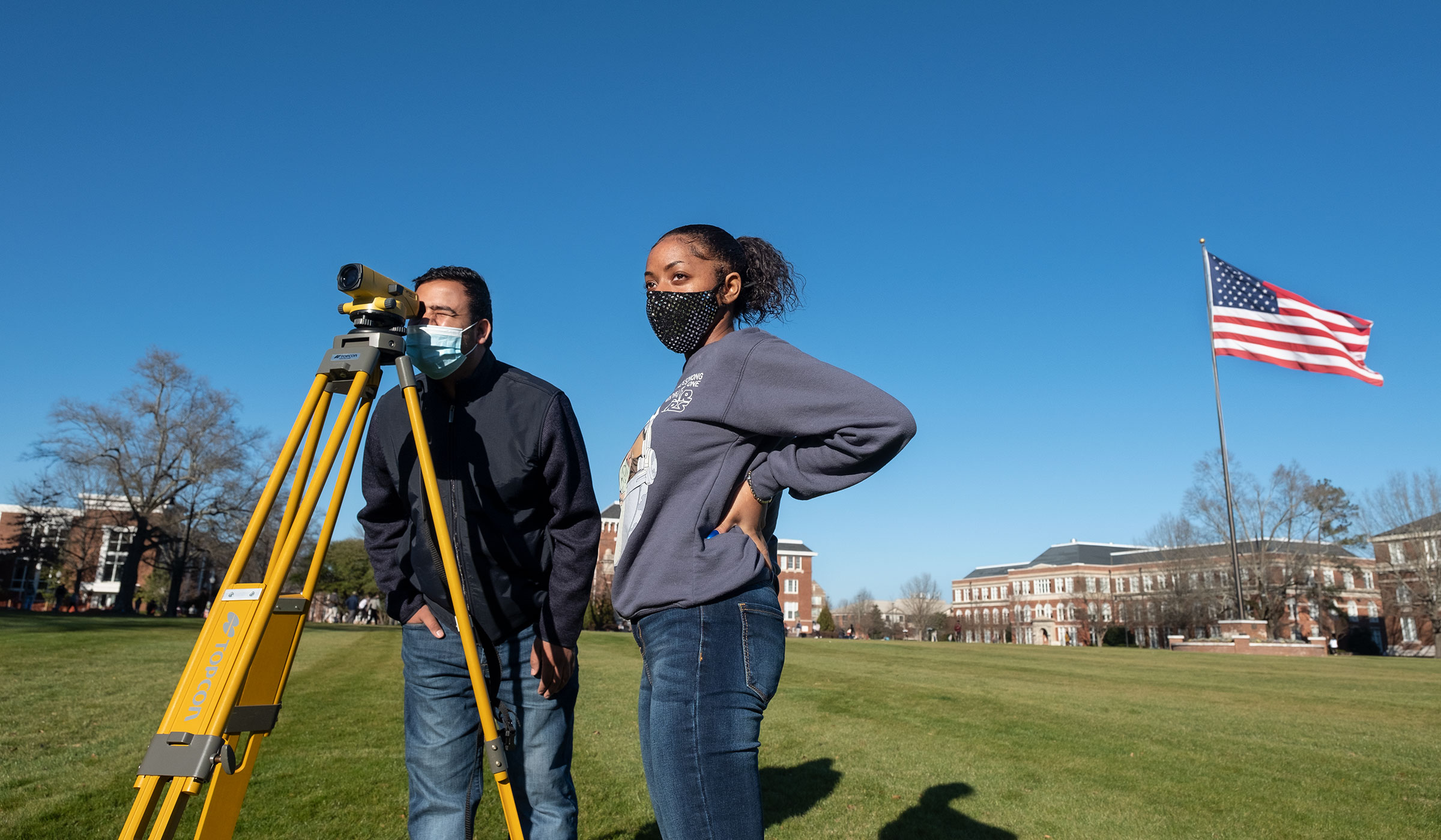 With a sunlit Drill Field in the background, Engineering student Nim Kc leans in to look through the survey viewfinder while Courtlyn Carson looks on.  Both wear masks for safety against Covid-19.