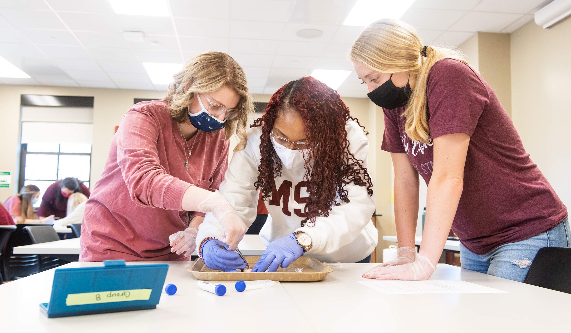 Female student in maroon, female student in white sweatshirt, and another female student in maroon leaning over tray on table