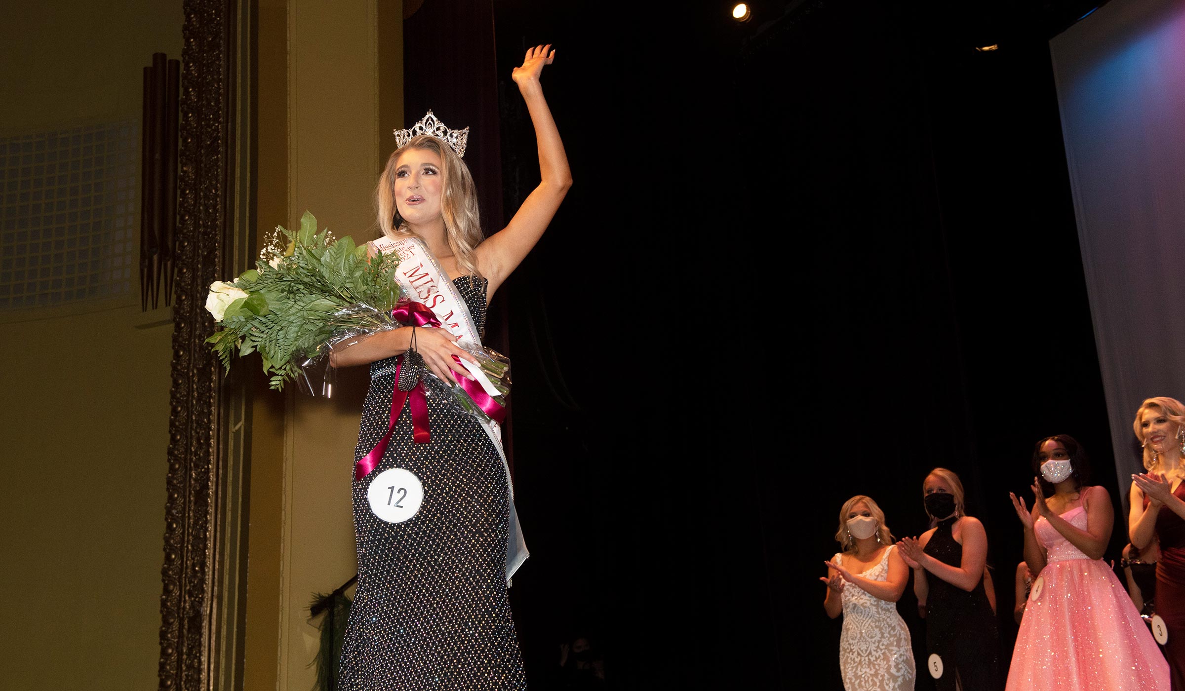 Woman in sparkly black dress, sash, and crown waving on stage