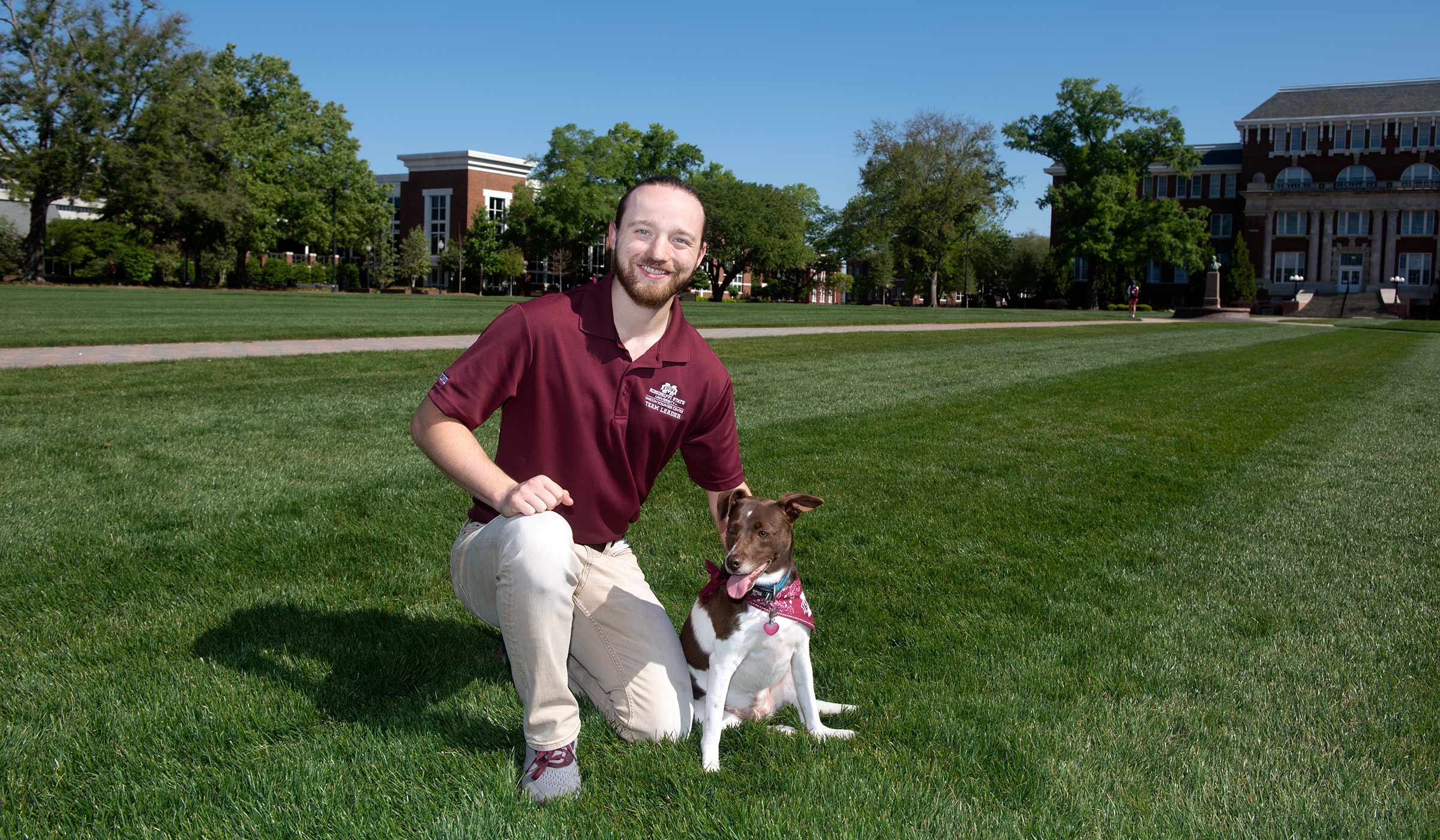Guy in maroon polo kneeling next to brown and white dog in grass on Drill Field