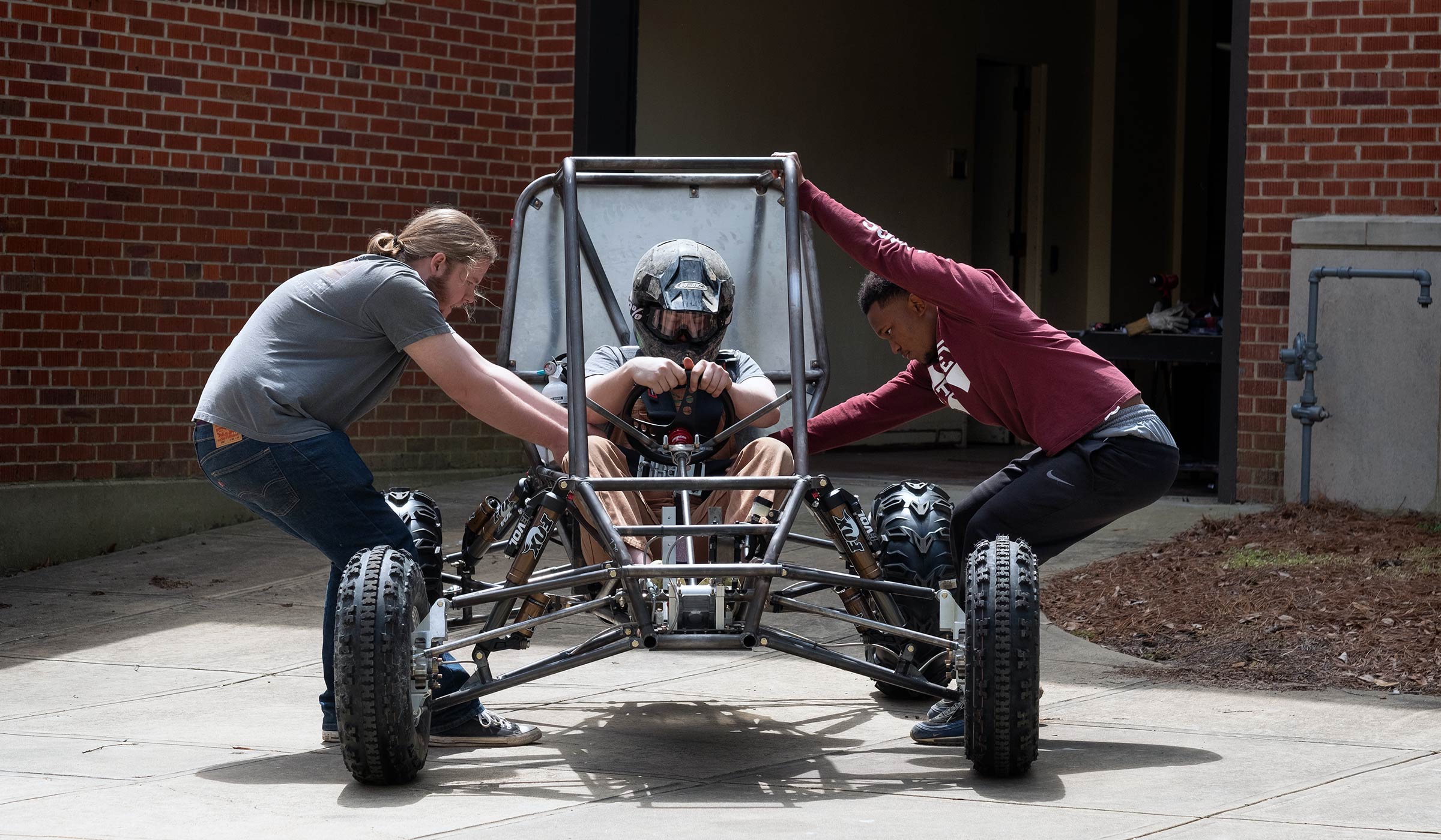 With a helmeted driver seated in the Baja car, two fellow Society of Automotive Engineers students pull on the seatbelt from either side to ready for the first drive.