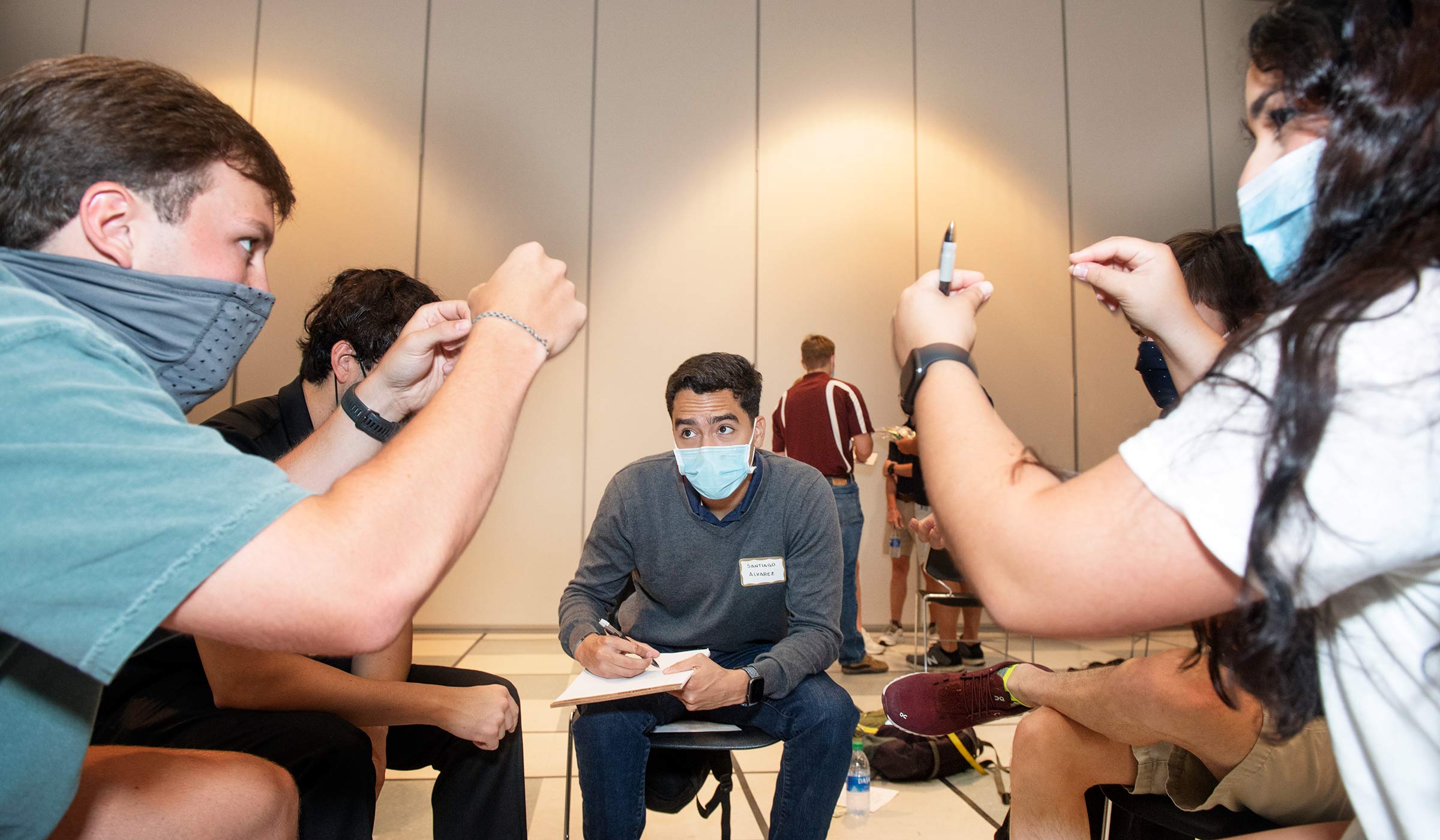 Masked students attempting to communicate an idea to onlooking student in gray sweater with notepad.