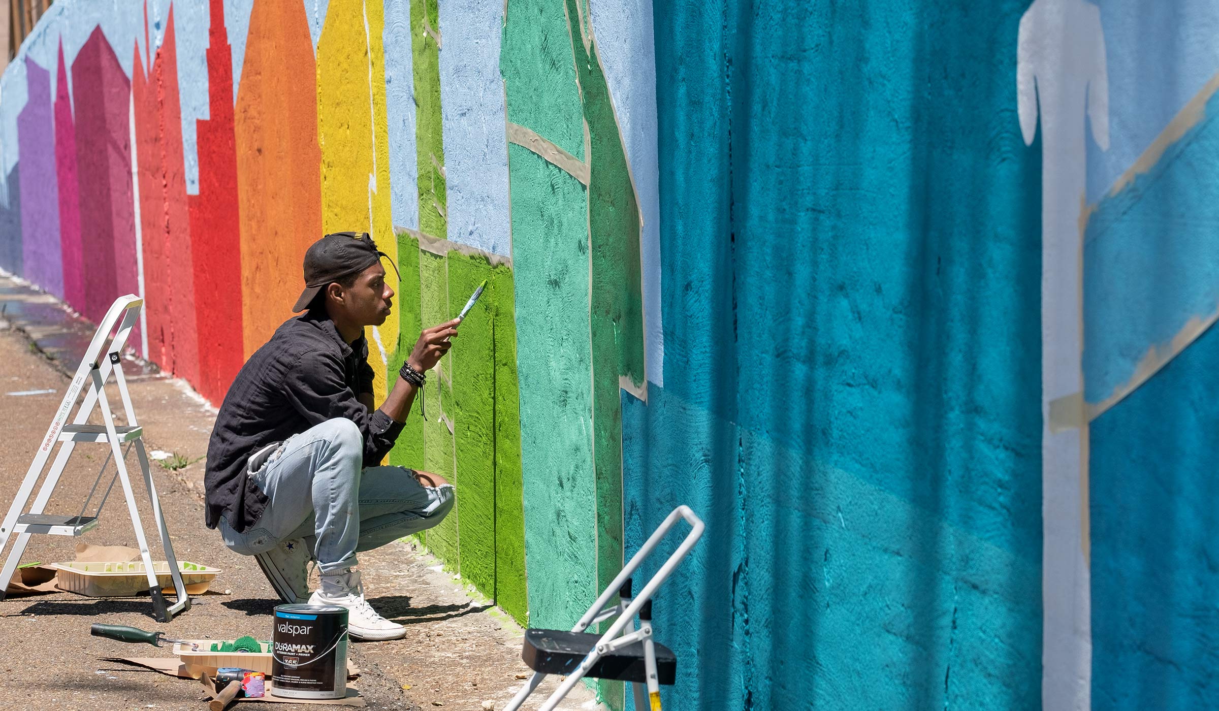 With the rainbow colors of buildings shapes painted on the wall mural to either side, a student uses a roller to apply green paint to his section.