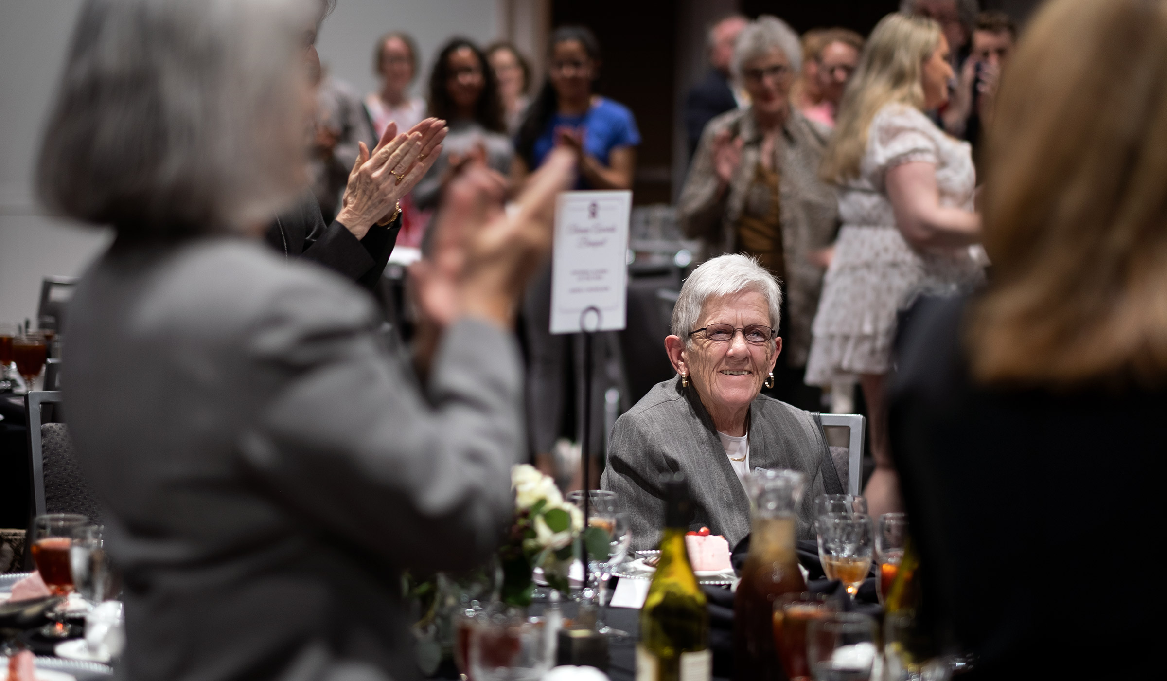 National Alumna of the Year, Janice Nicholson smiles while seated, as award banquet attendees stand and applaud her.