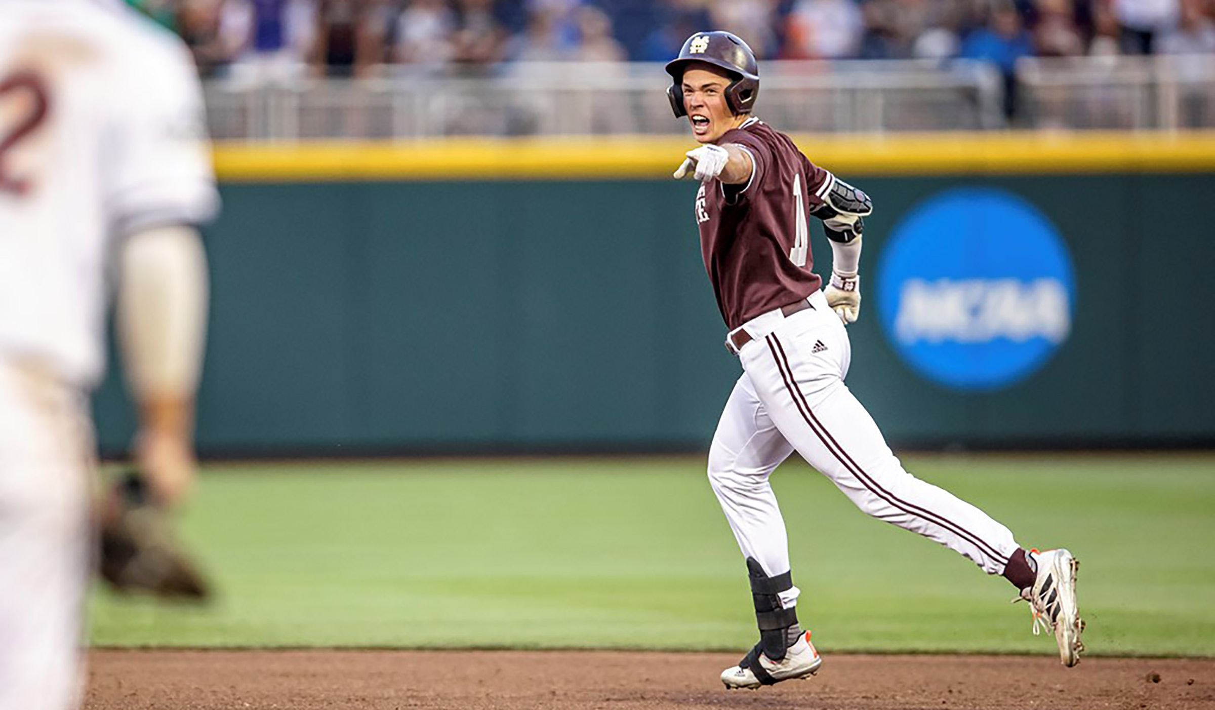 Guy in maroon baseball jersey and white baseball pants running bases while pointing