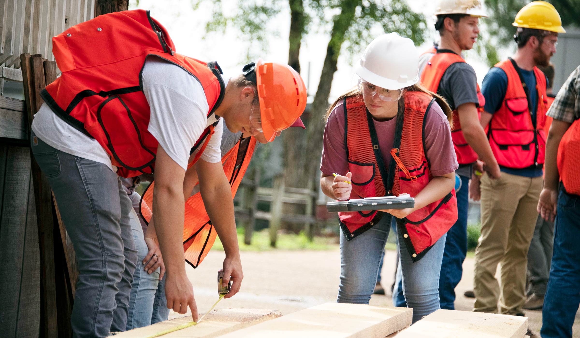 Guy in orange vest and hard hat measuring lumber while female in hard hat and vest takes down measurement
