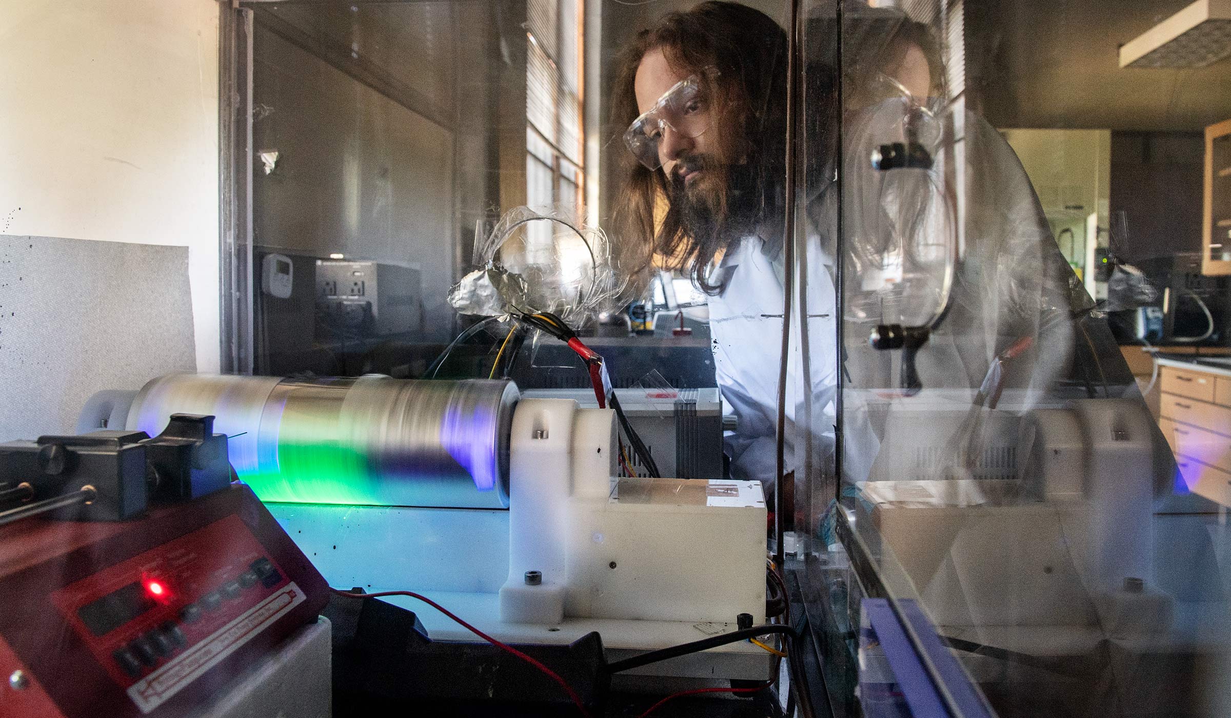 Wearing a white lab coat and protective glasses, REU student Ian Johnson looks in through the glass box on a glowing eletrospinning setup.