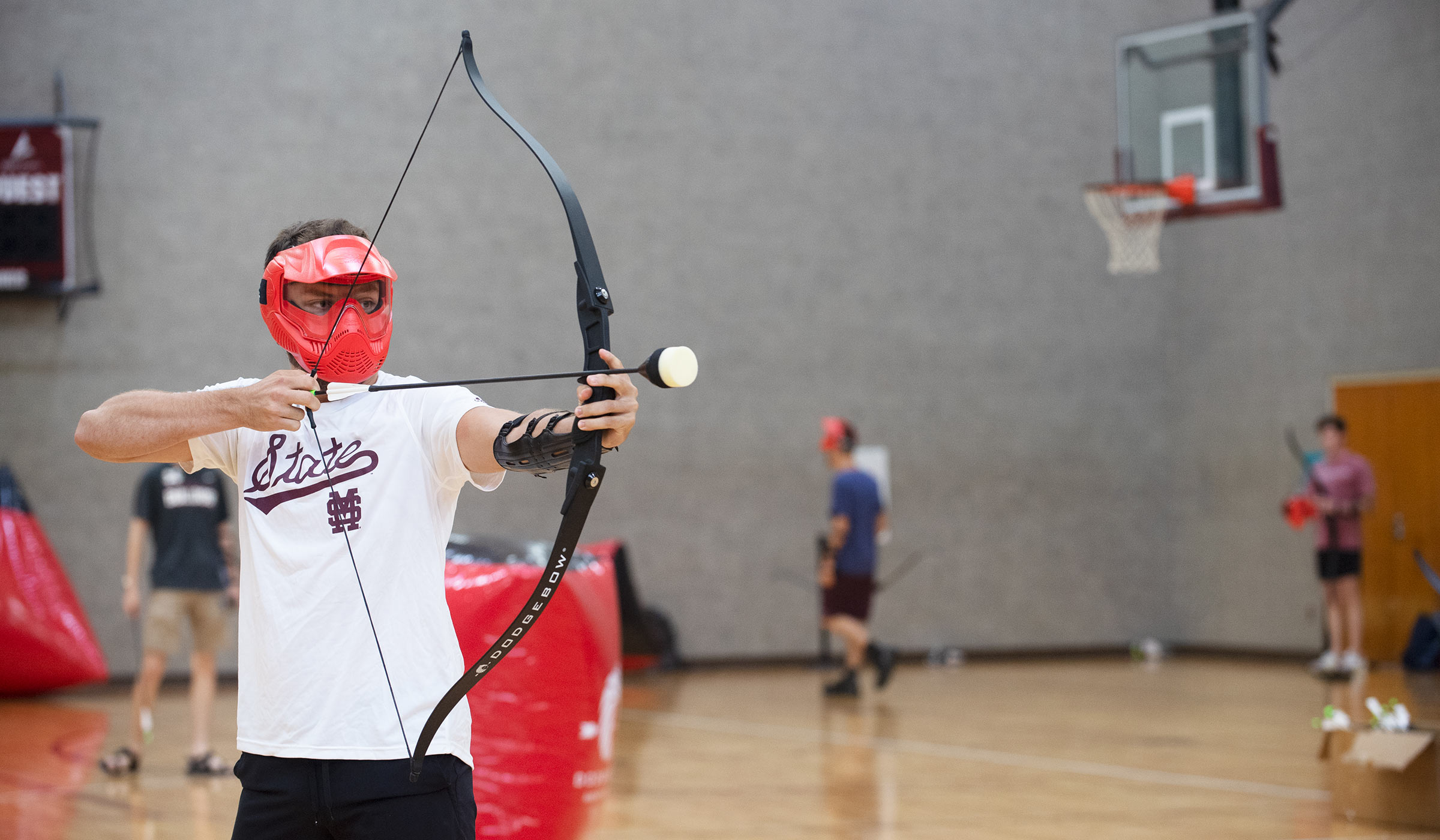 Student in helmet and white t-shirt with scripted state across front aiming foam arrow in gym
