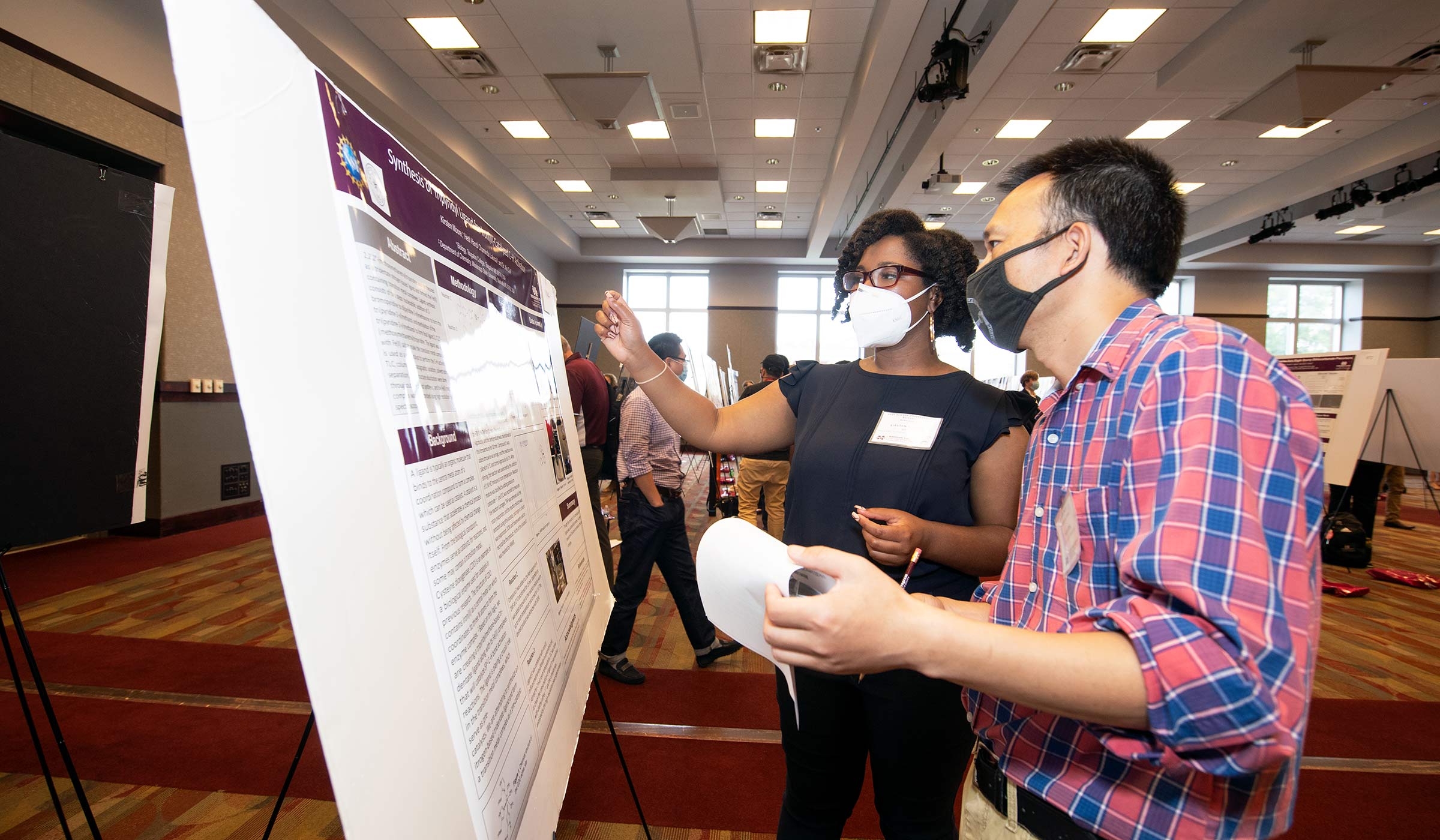 Female in dark attire and mask presenting research poster to a man in plaid shirt and mask