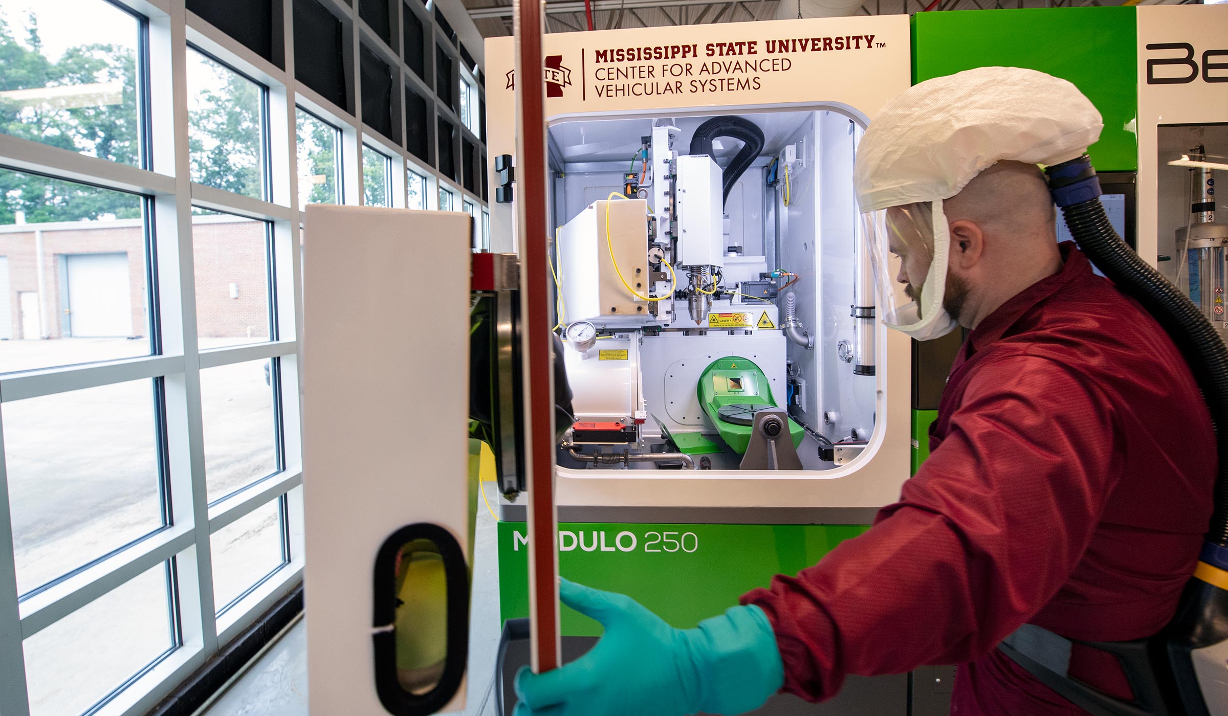 CAVS Research Engineer - wearing full face clear protective lab face covering - opens door to Additive Manufacturing Lab&#039;s large BeAM Modulo 250 machine, which is white and green.