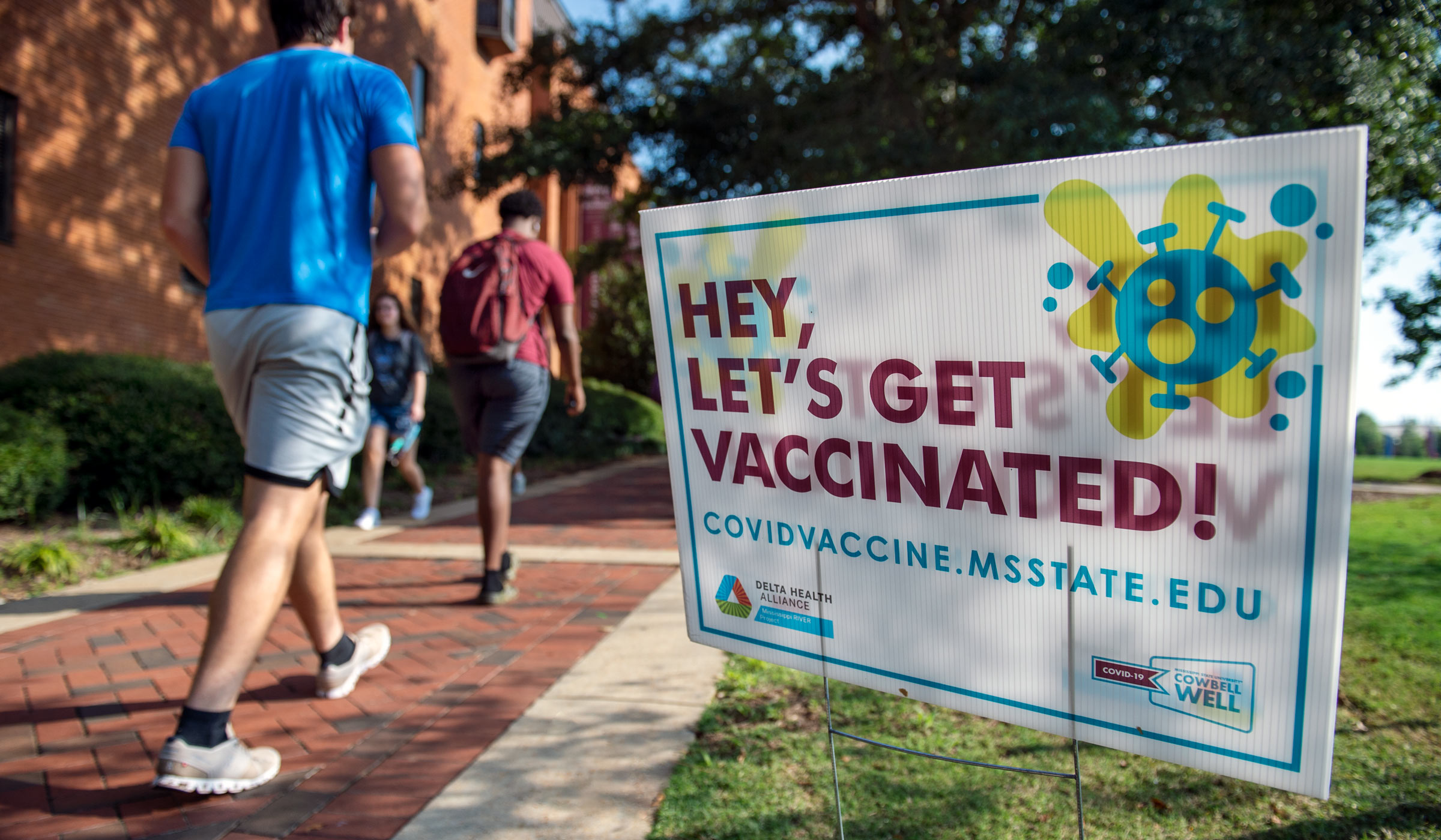 A sign encourages the campus community to &quot;Hey, Let&#039;s Get Vaccinated!&quot; with students walking past on a Drill Field sidewalk to the left.