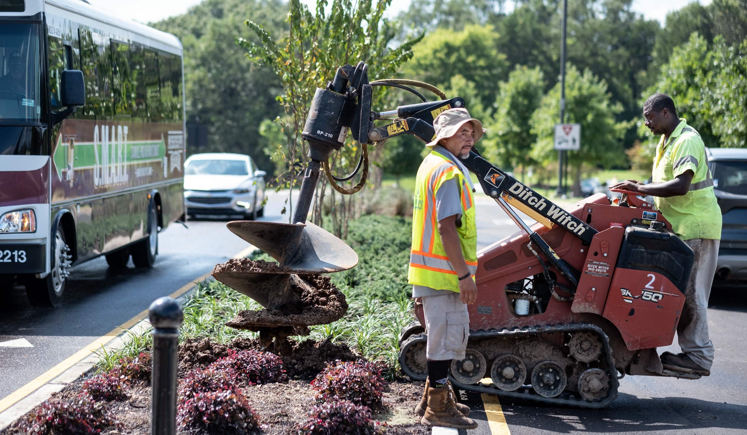Two members of the campus landscape crew work planting trees in the new landscaped median dividing Bailey Howell Drive. One worker operates a soil drilling machine, while a SMART shuttle bus passes to their left.