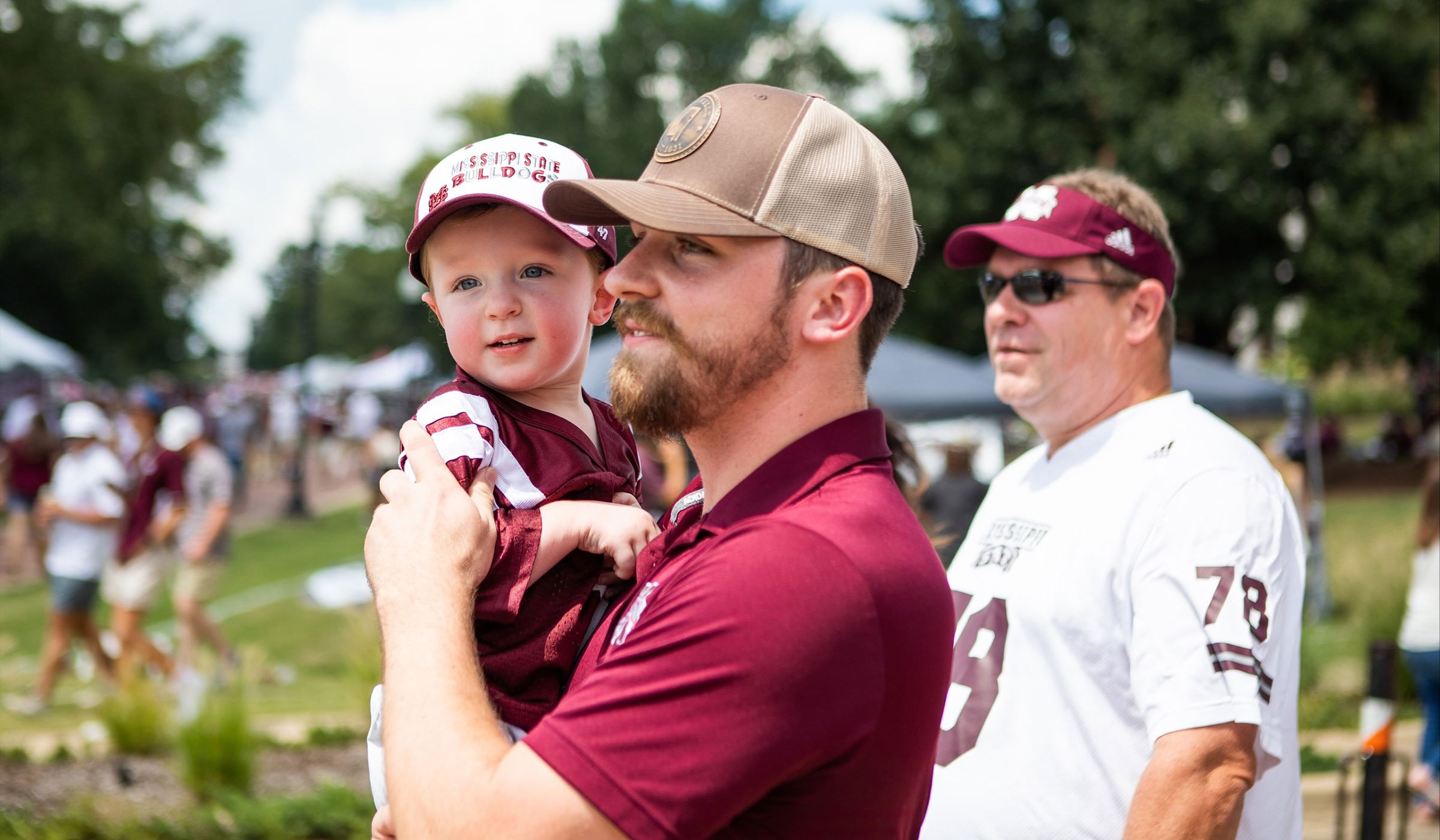 Man in maroon polo carrying kid in maroon and white jersey with baseball cap