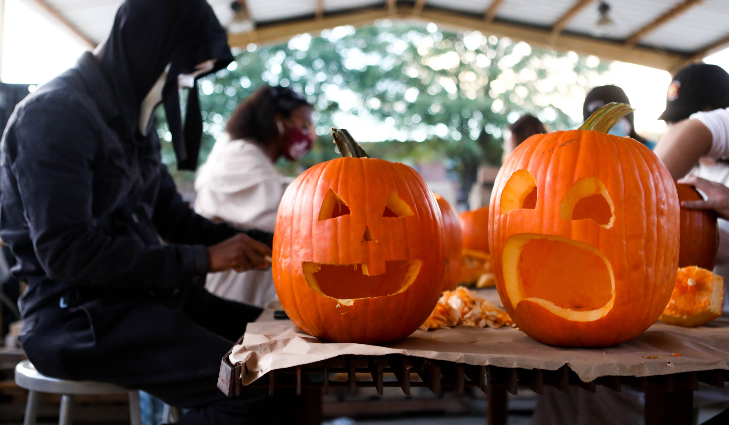 With two freshly carved Halloween pumpkins in the foreground, costumed students carve in the background, out of focus.
