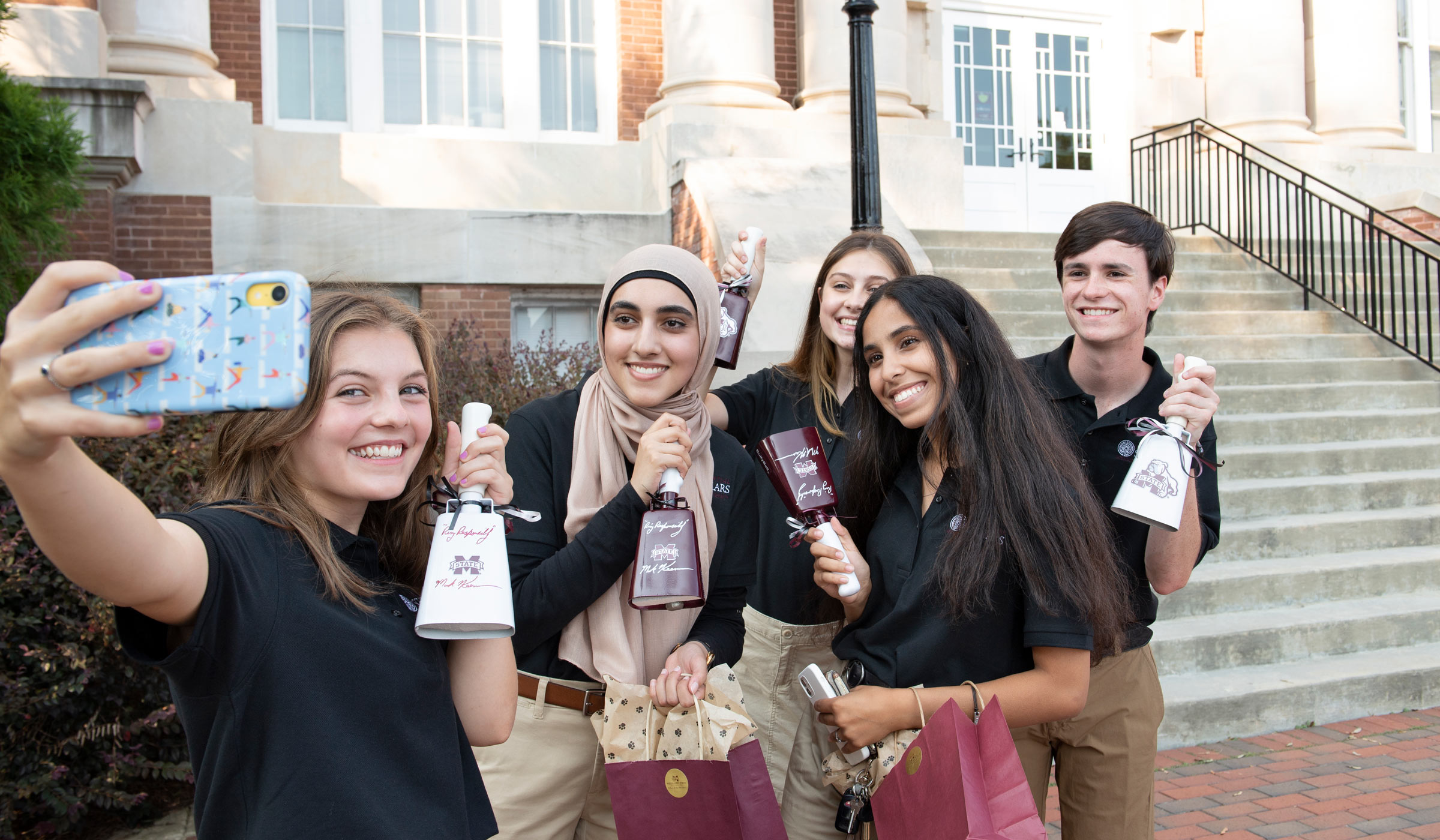 Holding a cell phone up on the left side of the frame, a female student takes a selfie with four fellow Presidential Scholars, each holding up their cowbell presents from President Keenum.