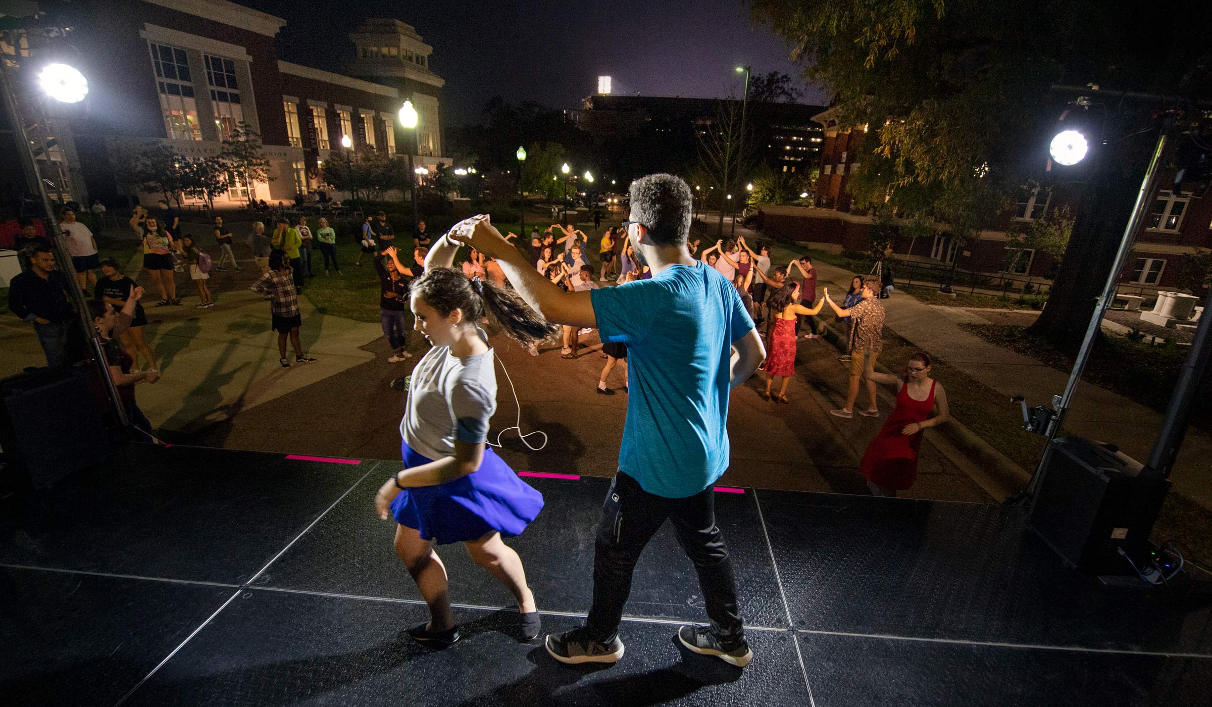 While teaching salsa to the attendees dancing in the street beyond, DJ twirls Samantha on the stage, lit by spotlights.