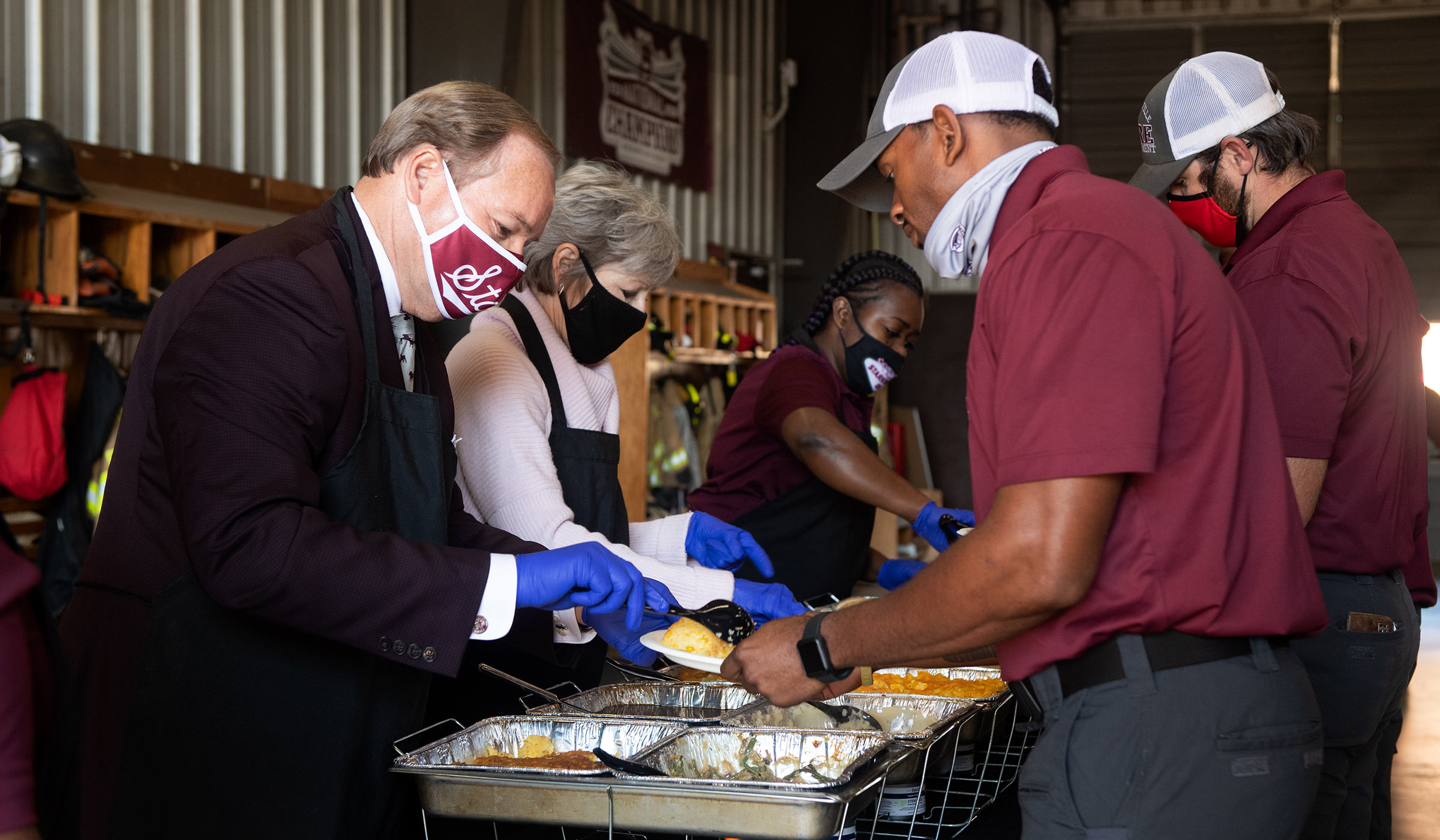 Man in maroon blazer and mask serving food to man in maroon shirt at fire station