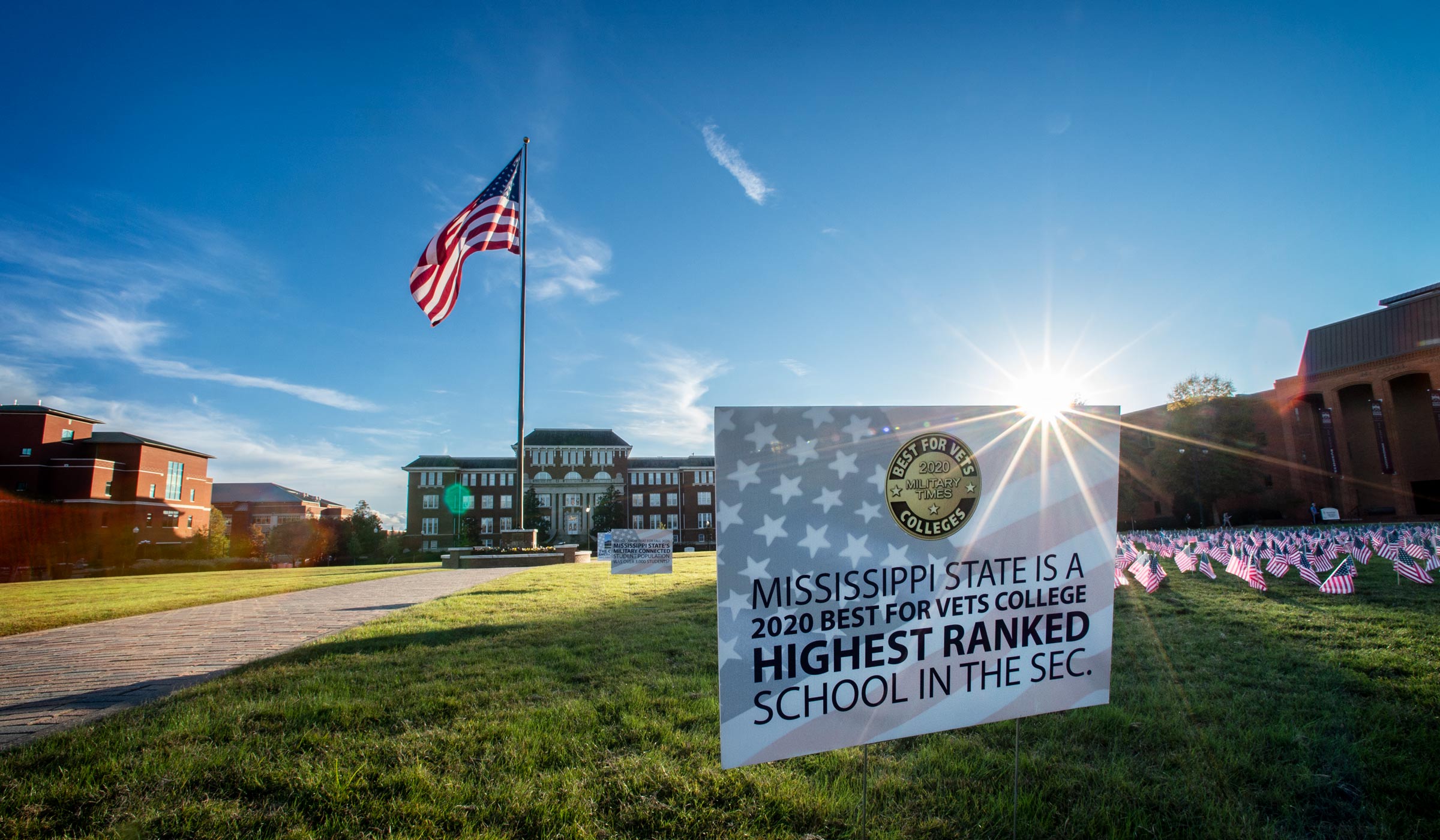 Sunflare backlights sign on Drill Field next to Veterans Day American flags: MSU is a &quot;2020 Best for Vets College Highest Ranked School in the SEC.&quot;