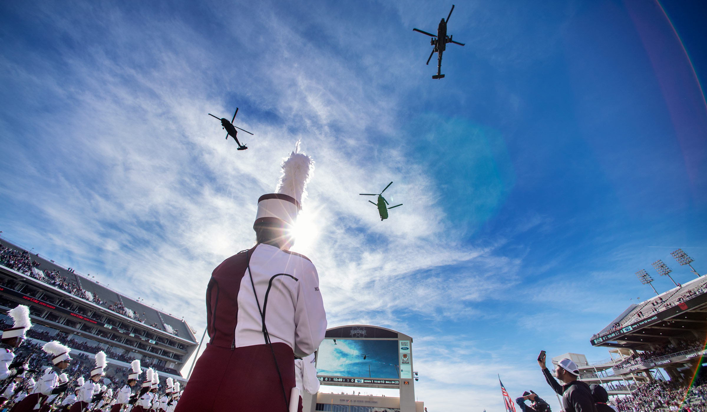 Viewed looking up from below MSU Band members on the Dudy Noble Field, three Blackhawk helicoptors flyover the stadium, with blue sky beyond.