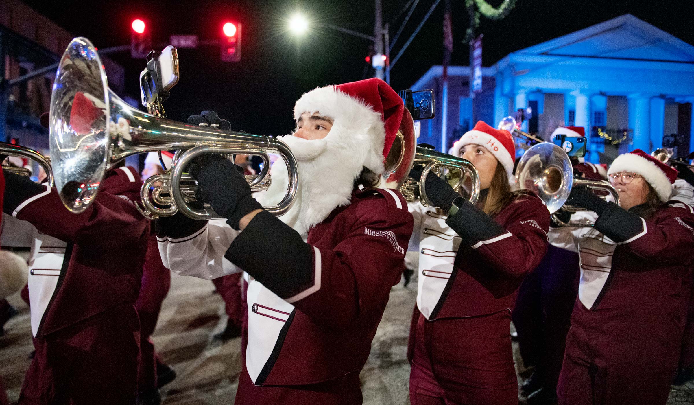 Wearing a Santa hat and white beard, a band member marches past Starkville downtown&#039;s courthouse at night with his fellow mellophone players.