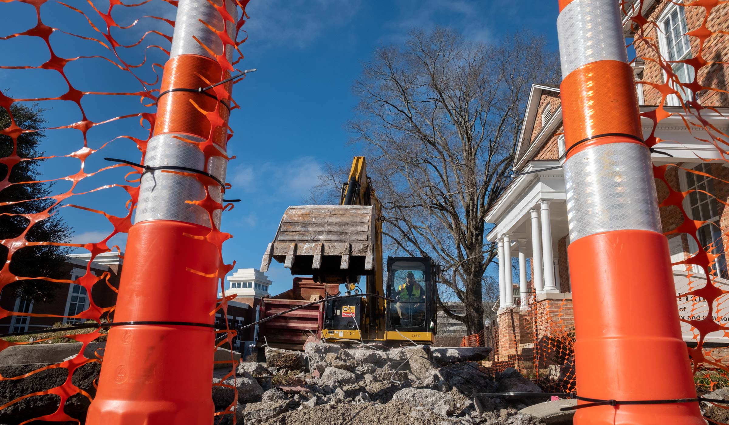 Bright orange construction cones frame a bulldozer tearing up the concrete sidewalk in front of George Hall with a blue sky beyond.