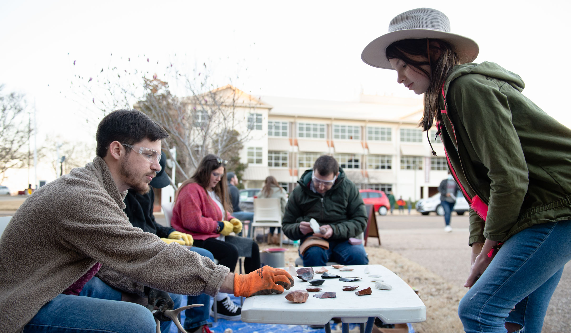An Anthropology graduate student wearing safely glasses pauses in his work on antlers to discuss the flint and obsidian samples on a table with a hat-wearing girl visitor.