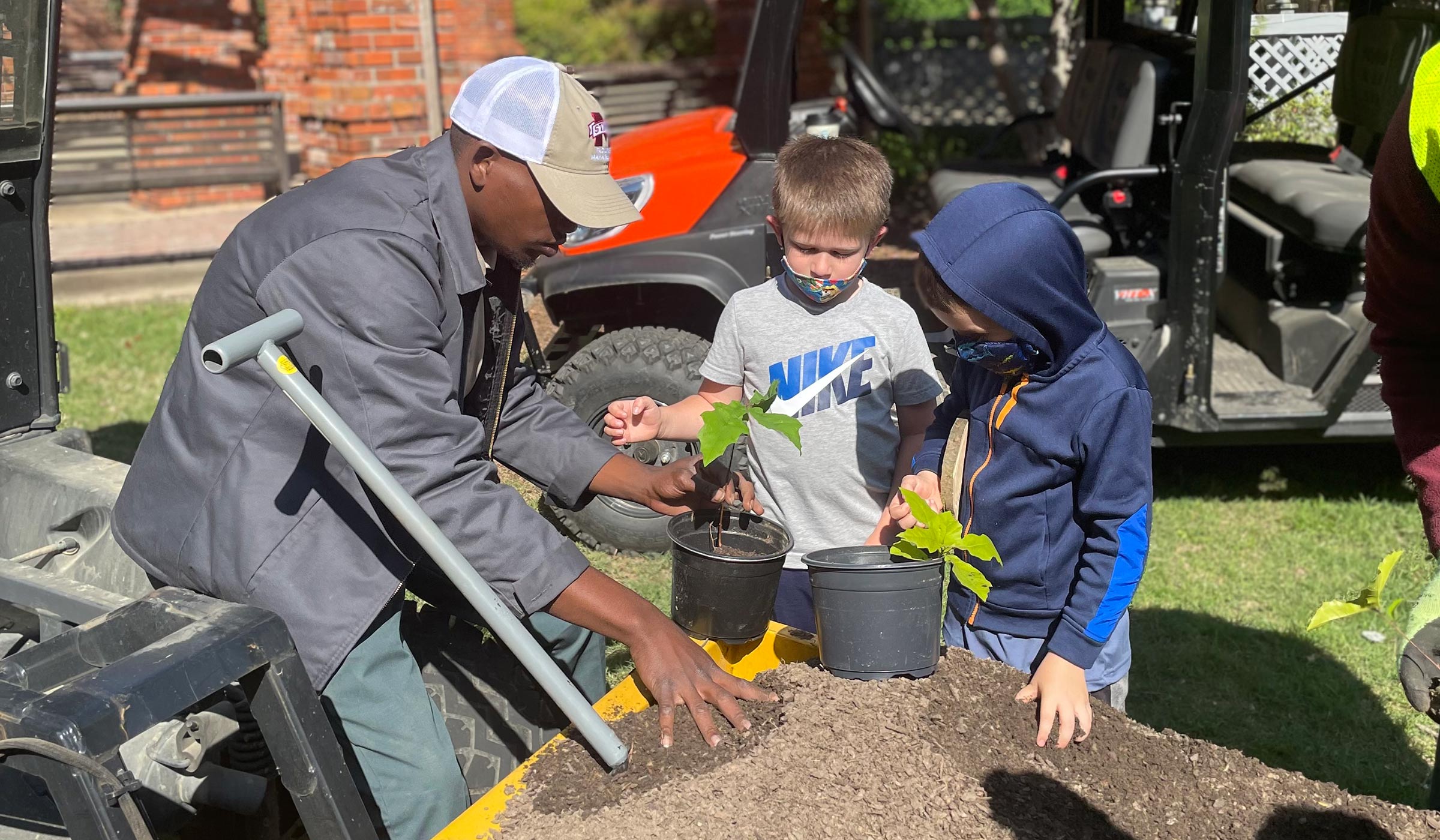Guy in gray jacket and cap helping two kids add soil to tree clippings in black pot.