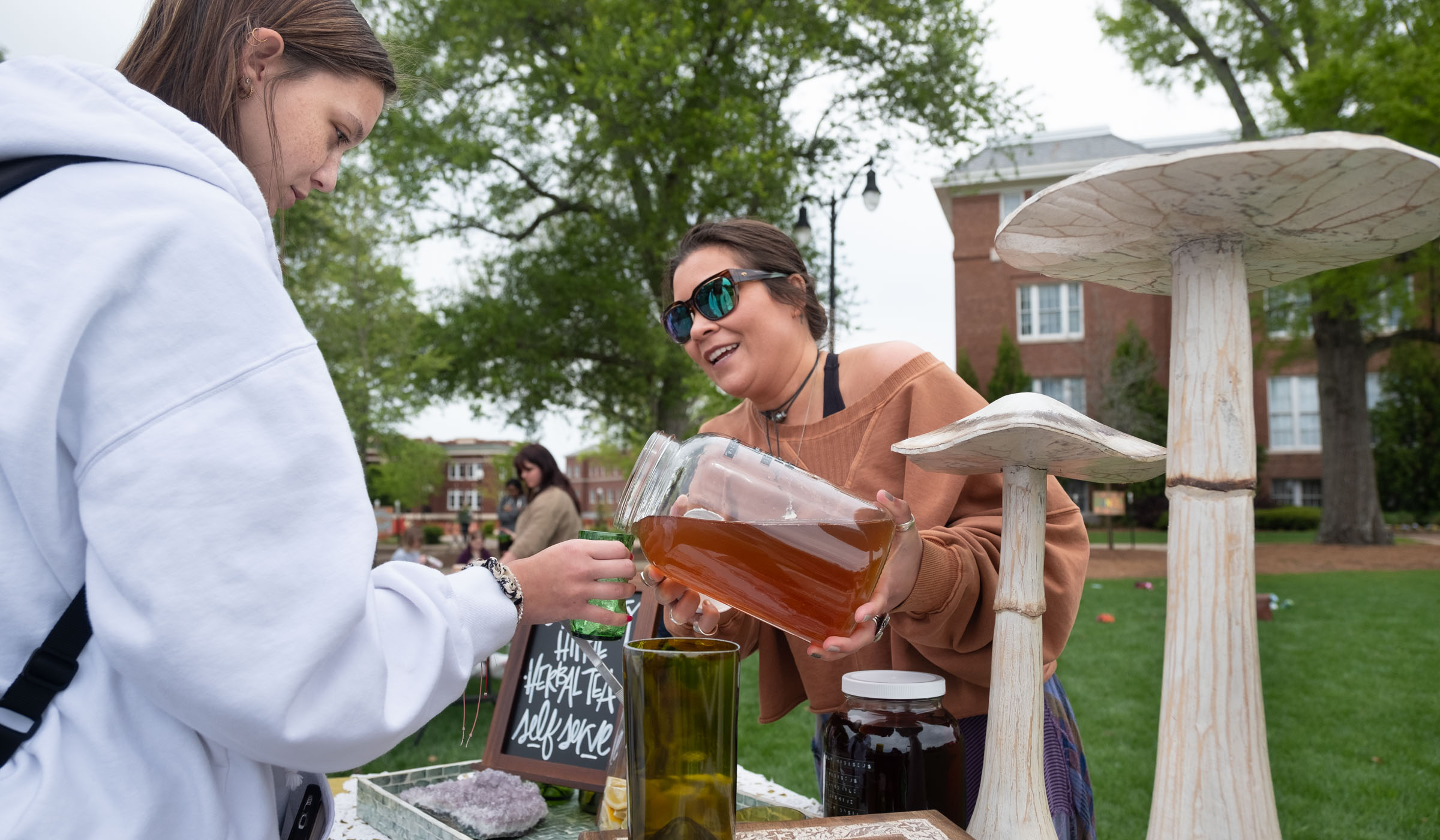 With large mushroom sculptures to the right of the frame, a student receives tea poured into a glass by a booth vendor.l