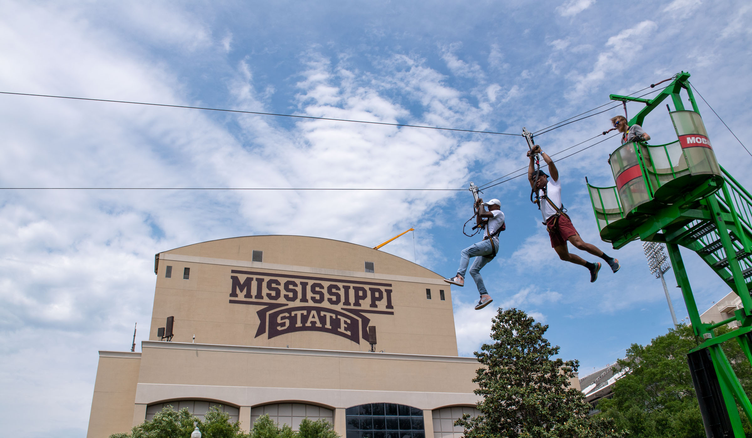 Two students launch down a zipline at the Junction, with the Mississippi State logo of the Jumbotron behind them.