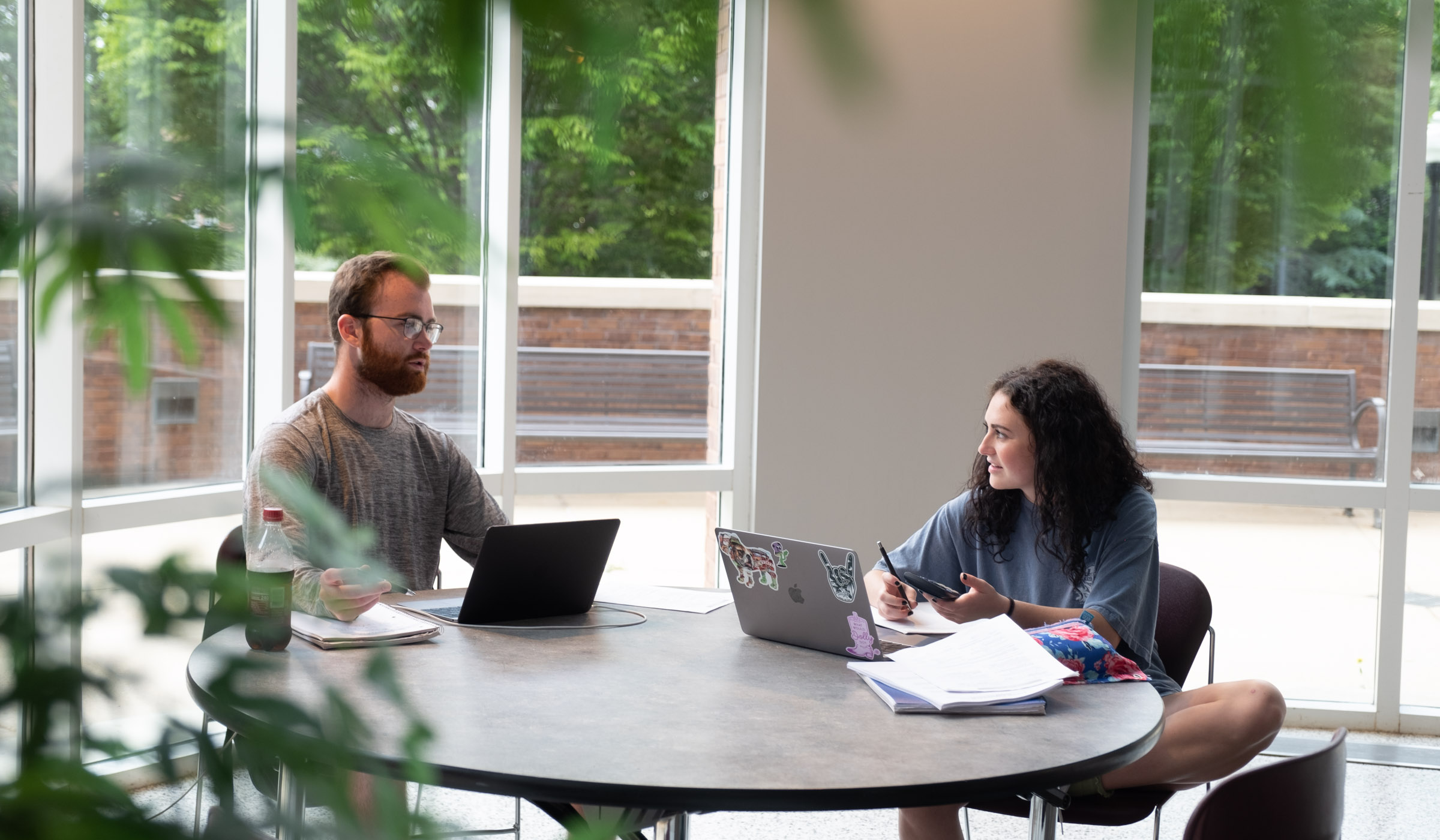 Two students study near the windows of McCool&#039;s Atrium, working at laptops at a round table.