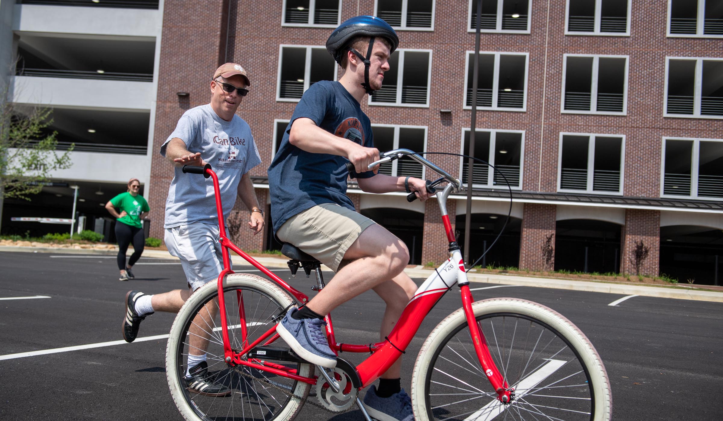 iCanBike Camp participant Carter Oswalt rides a red bike in a sunny parking lot next to Parking Garage North while Camp Director Twietmeyer runs next to him, ready to catch him as needed.