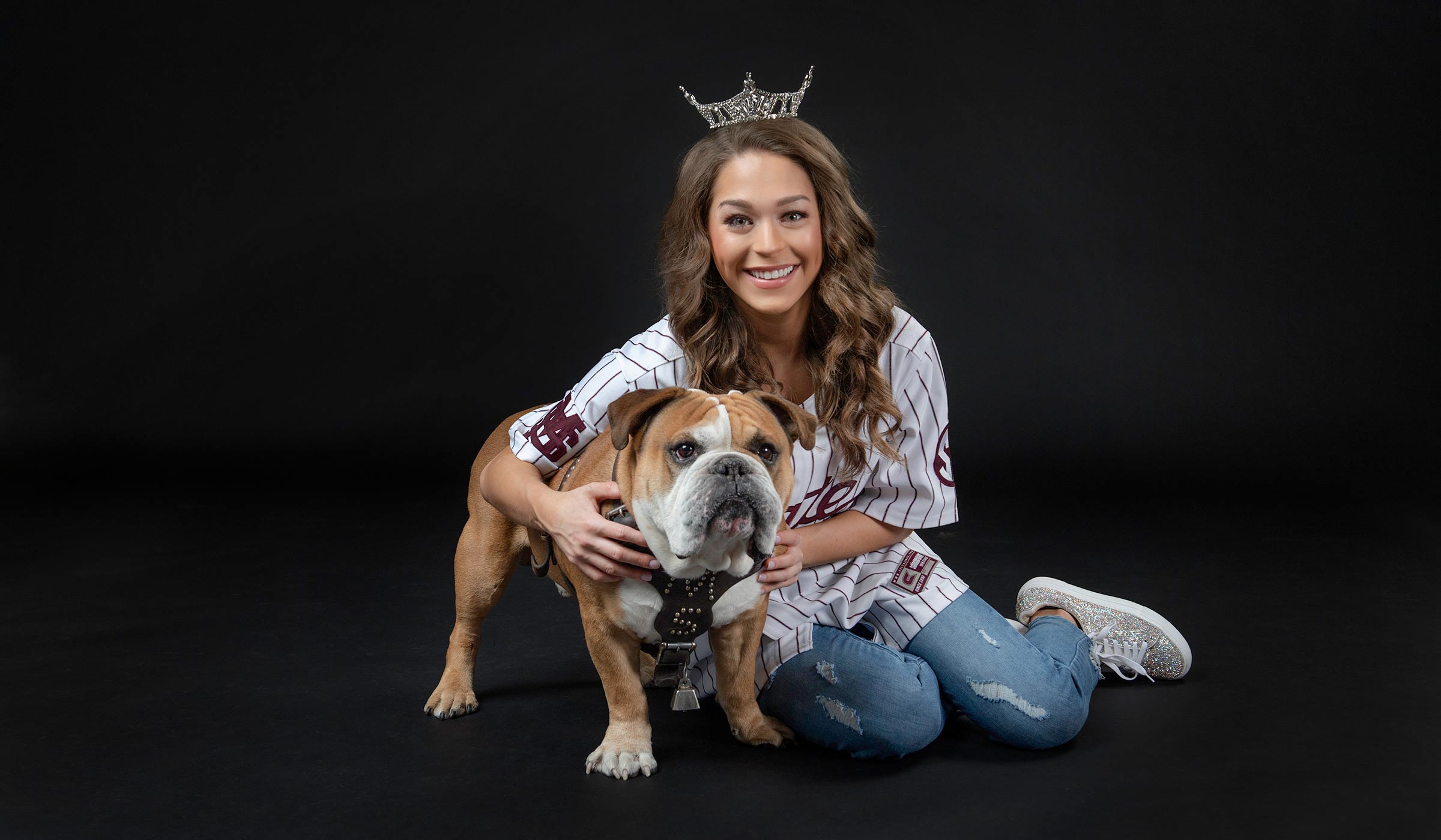 Girl in crown and baseball jersey sitting next to bulldog in studio