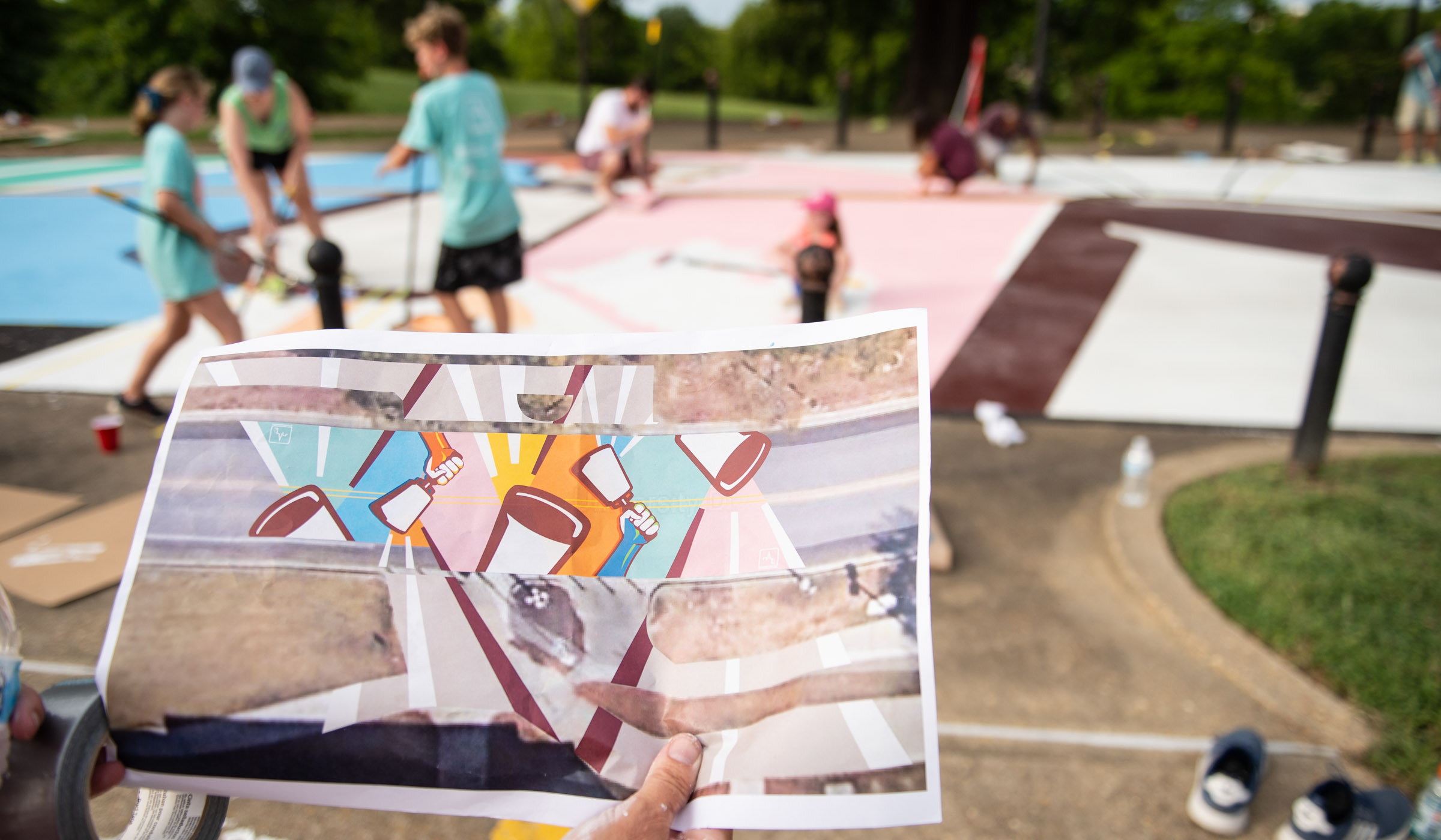 A hand holds an illustration of the finished product look of thes treet mural, with people painting the street in the background.