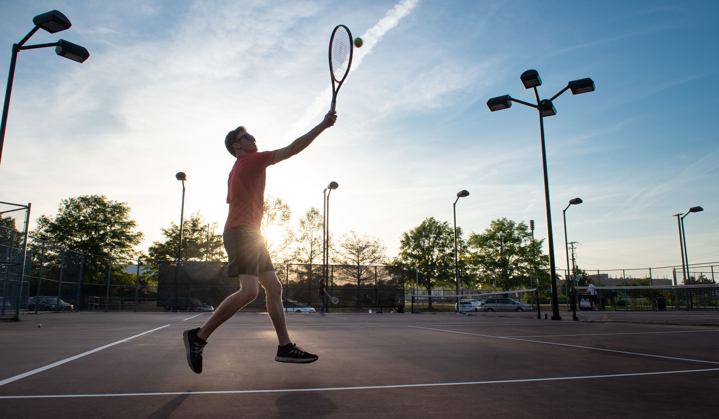 With the setting sun behind him, a tennis player leaps to hit a ball on the Sawyer Tennis Courts.