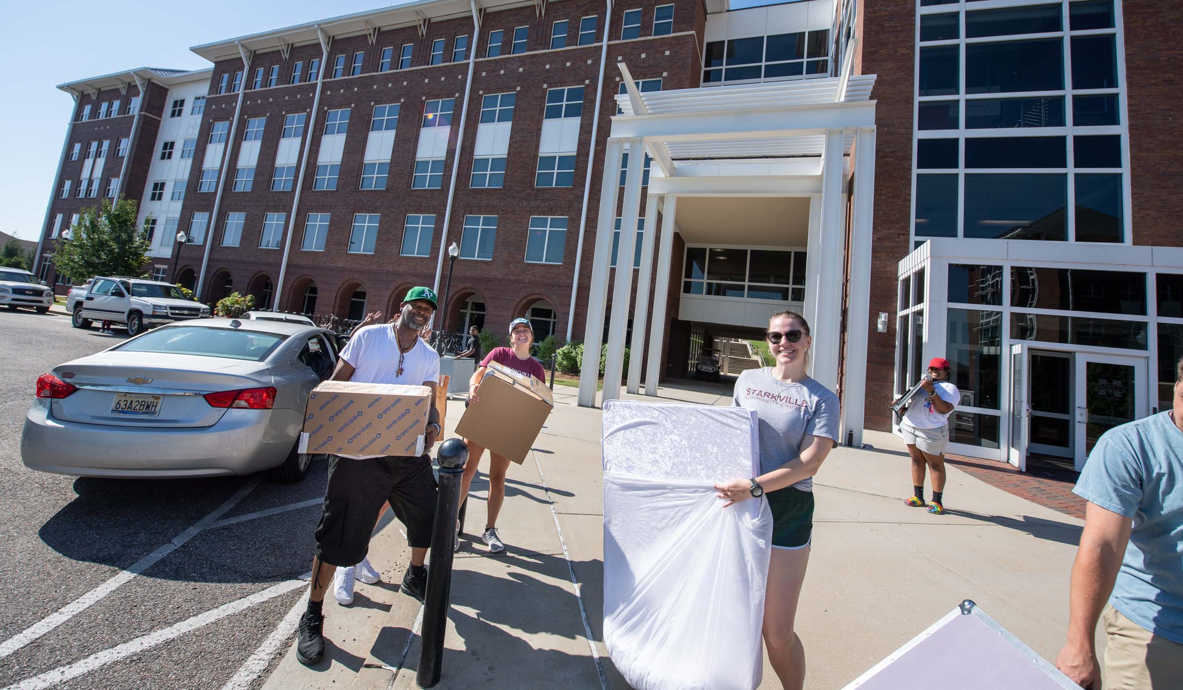 A msattering of five people stand holding items moving into Dogwood Hall on Move-In Day, with the residence hall behind them.