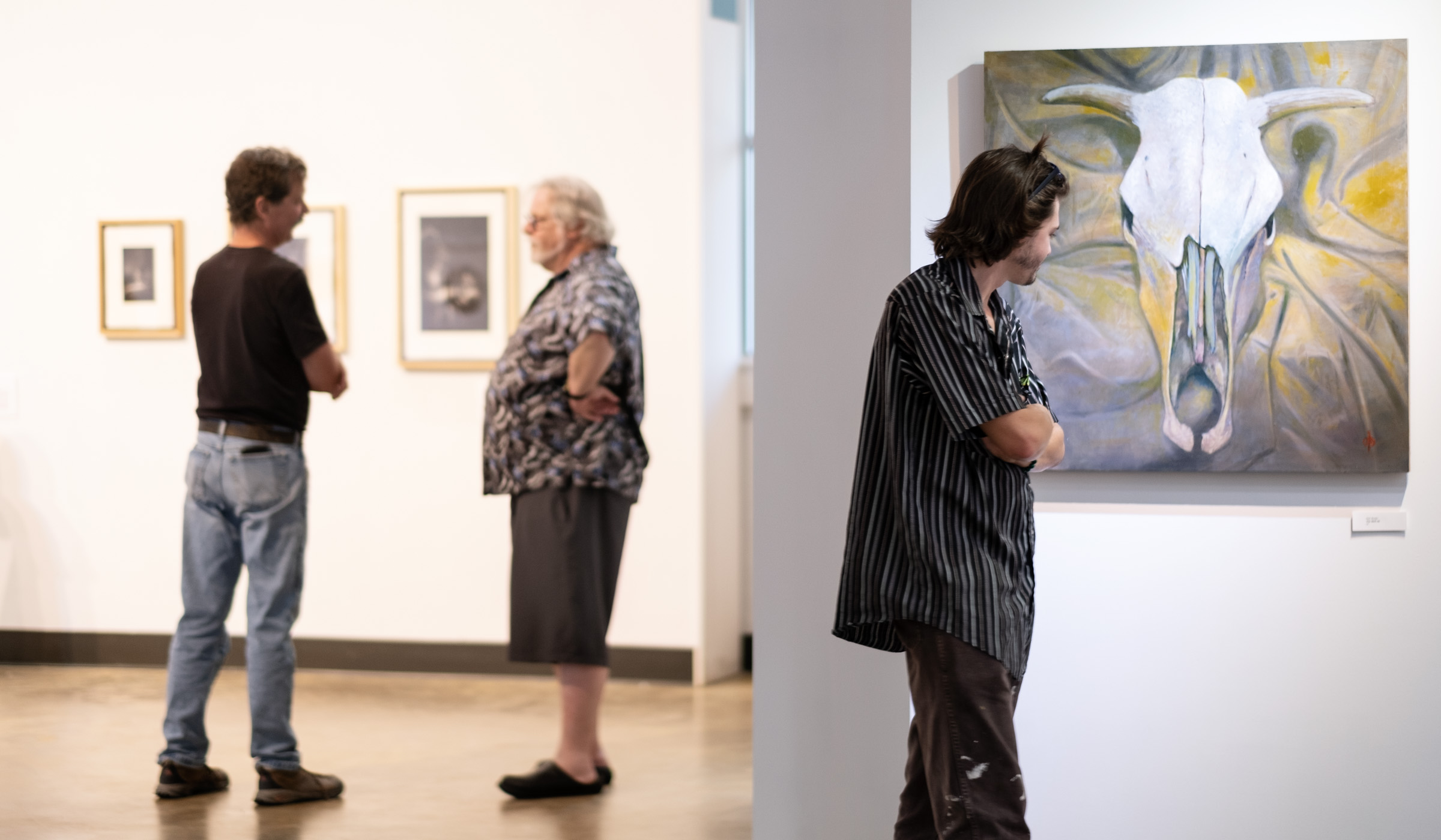 A male visitor to the art gallery looks at a painting of a cow skull in the foreground while two other vistors chat in the background.