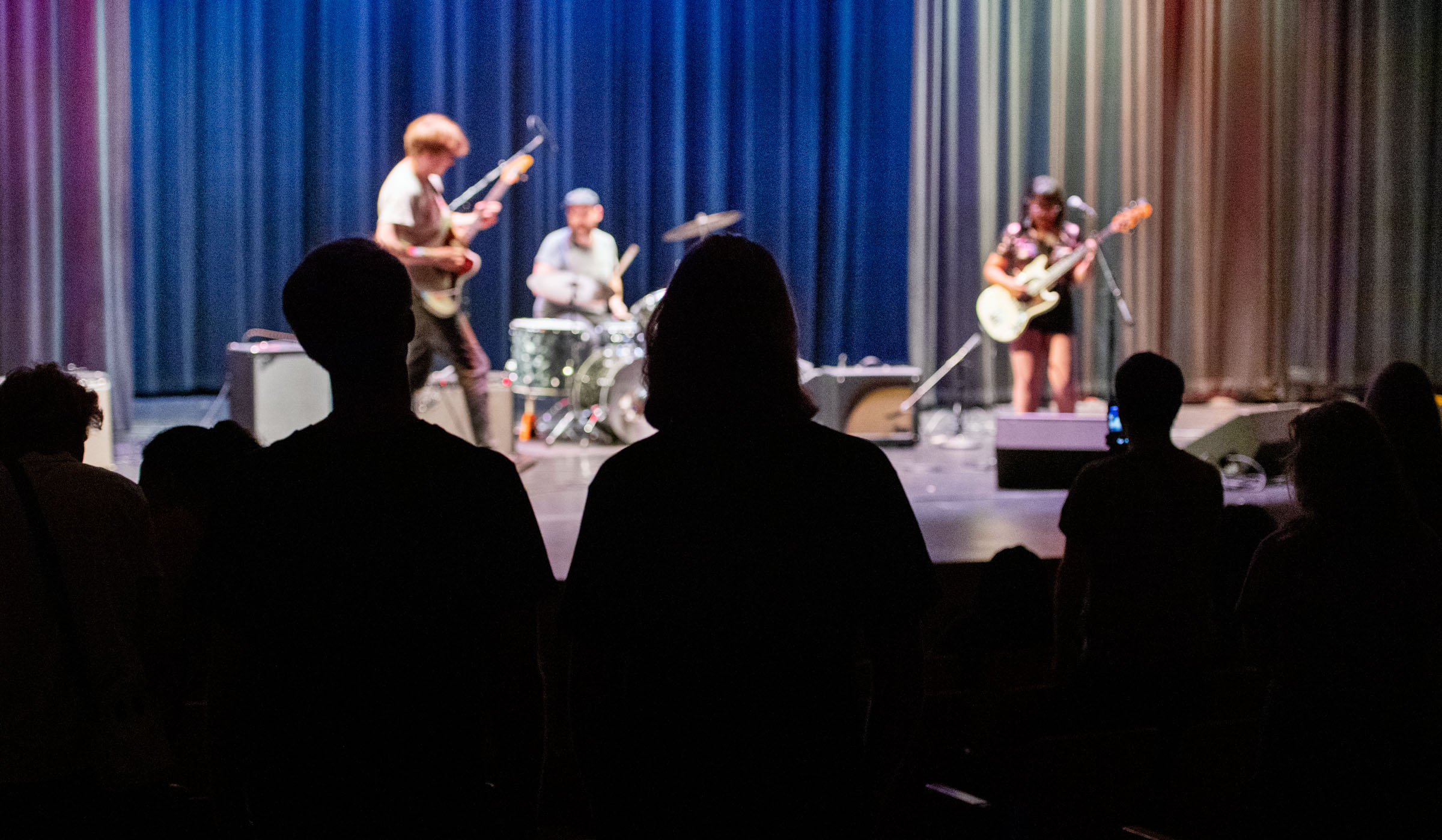 With dark sillouettes of concert goers in the foreground, a three piece rock band is lit up on the stage beyond.