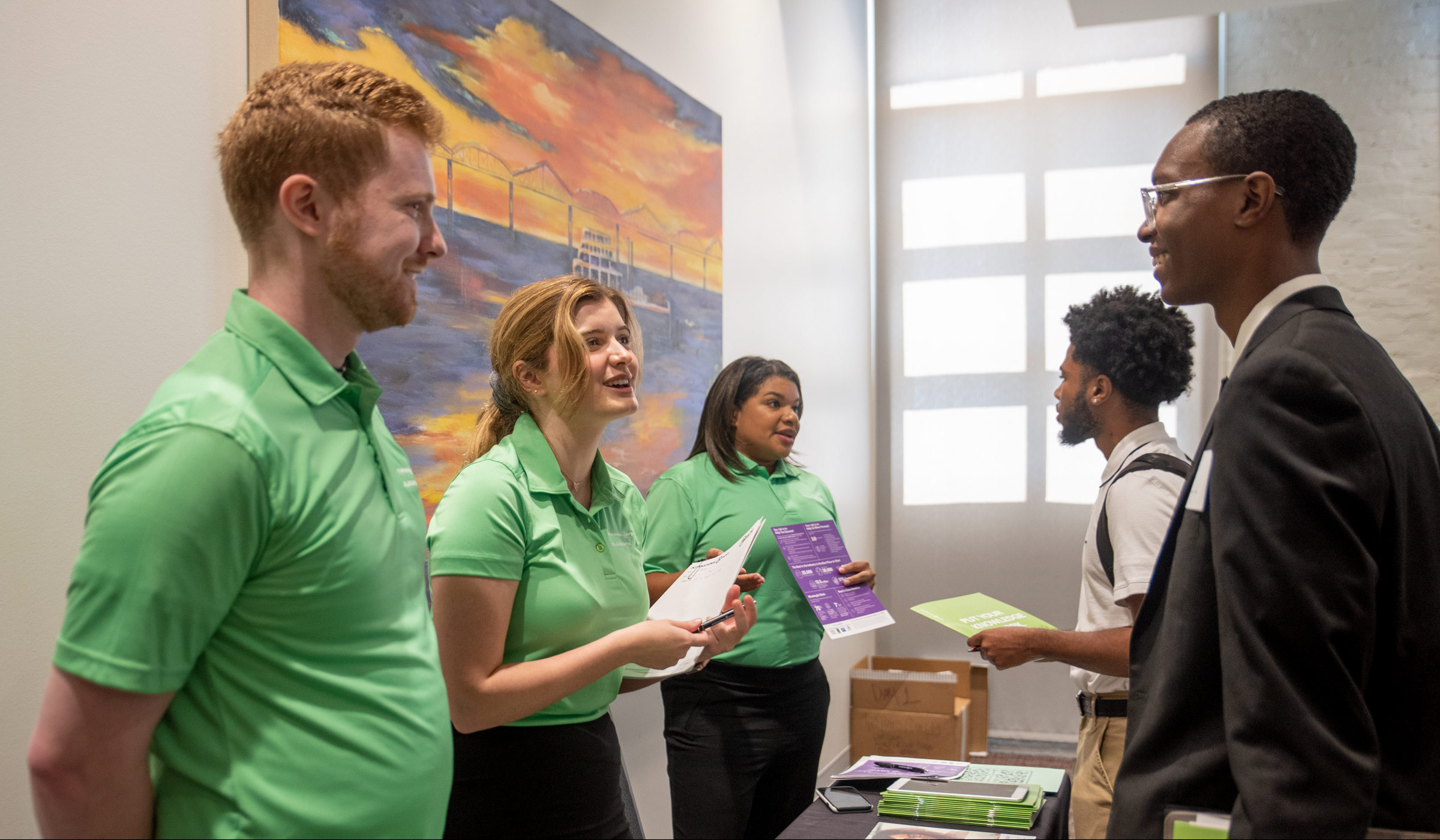 Three employer representatives wearing green shirts talk about their company with two students wearing suits.