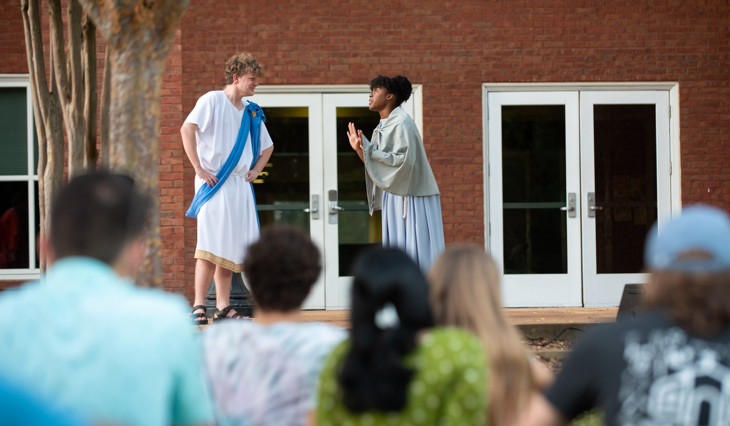 Two student actors in togas peform outside Griffis Hall doors, with the backs of audience members in the foreground.