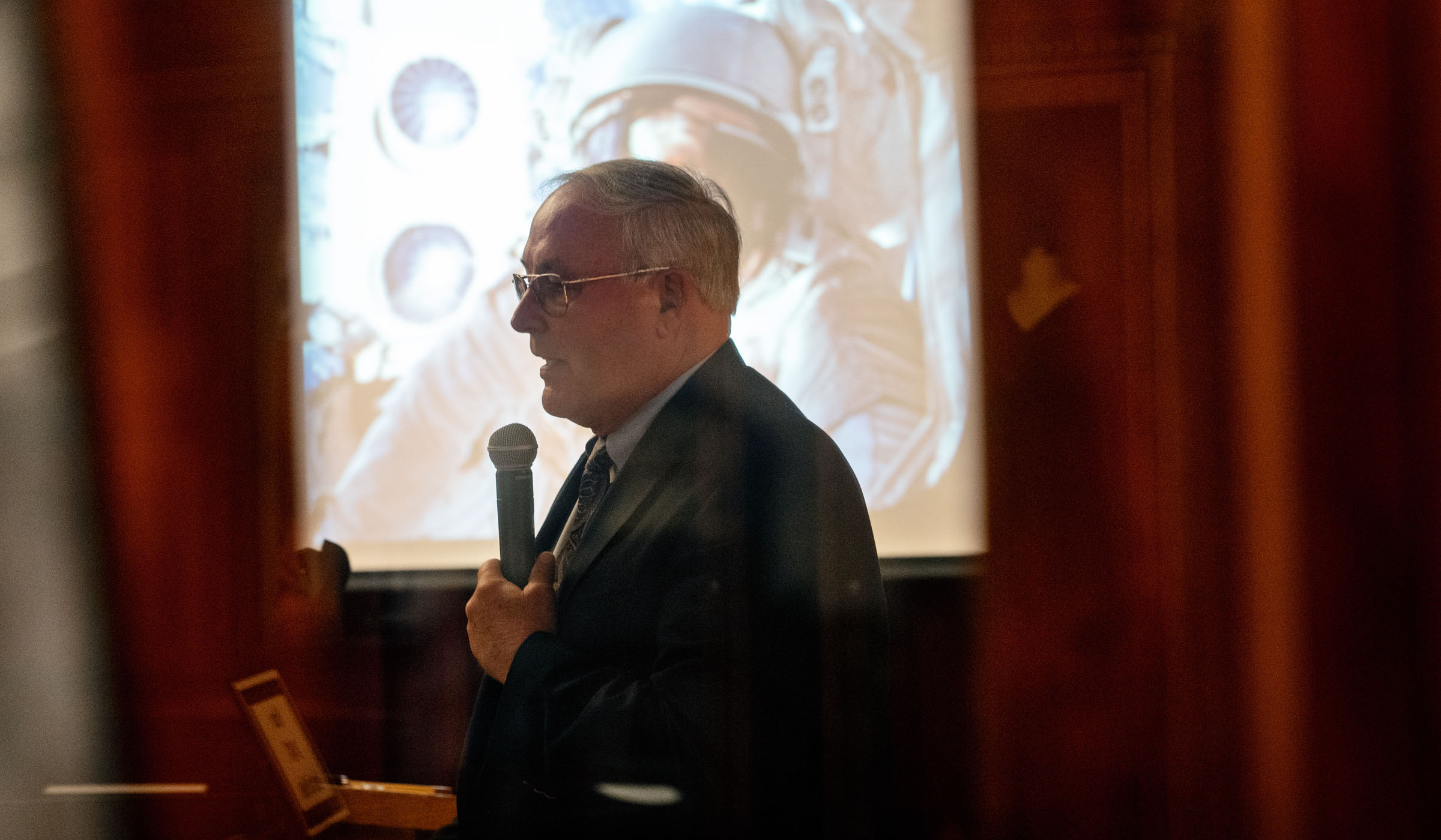 NASA Astronaut is silhouetted in profile, speaking in front of a photo of himself on one of his seven shuttle space flights. 