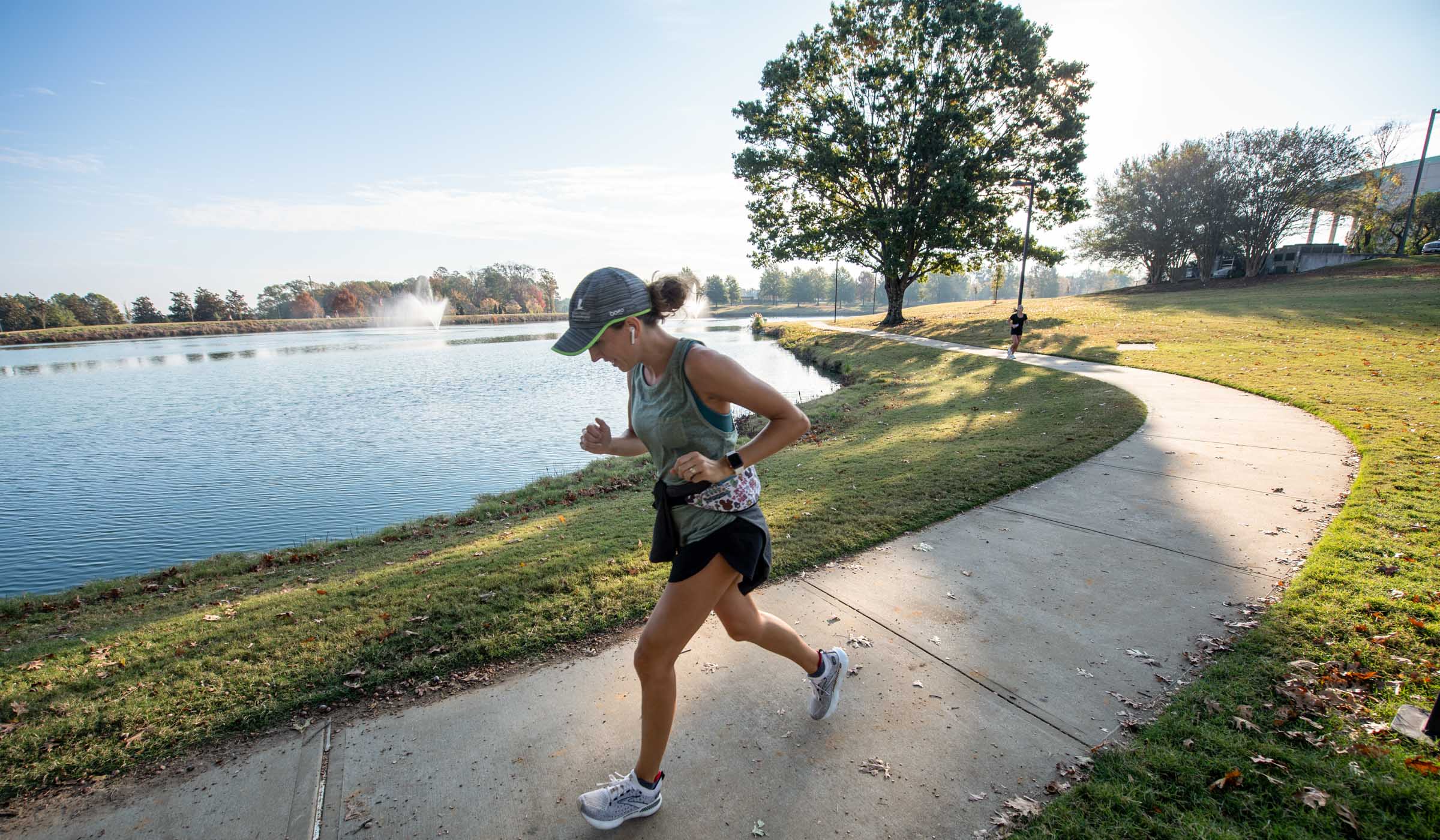 A female jogger runs on the sidewalk in the foreground, with Chadwick Lake and a tree in the background.