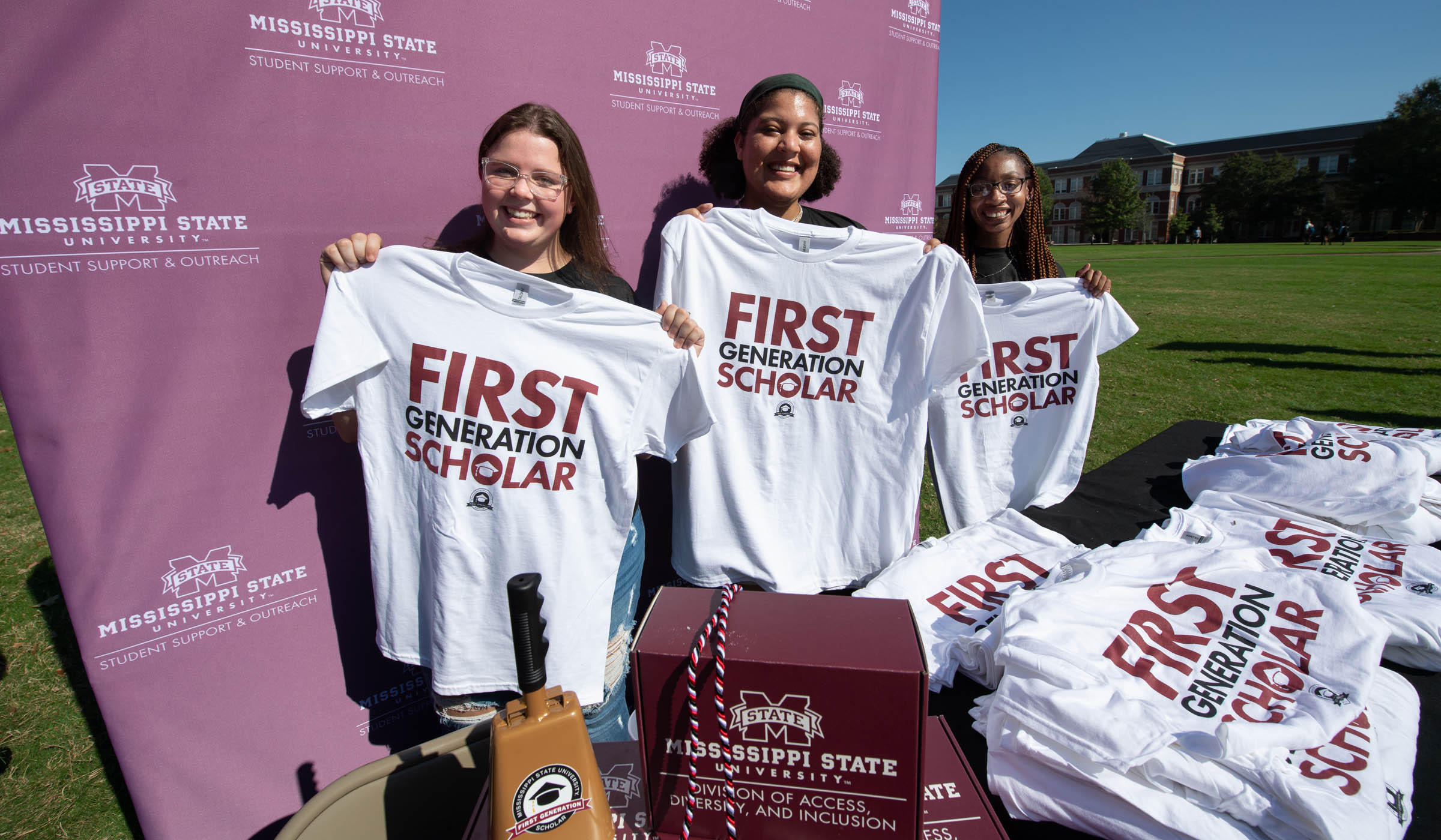 Three students hold up First Generation Scholar t-shirts at their Drill Field table.