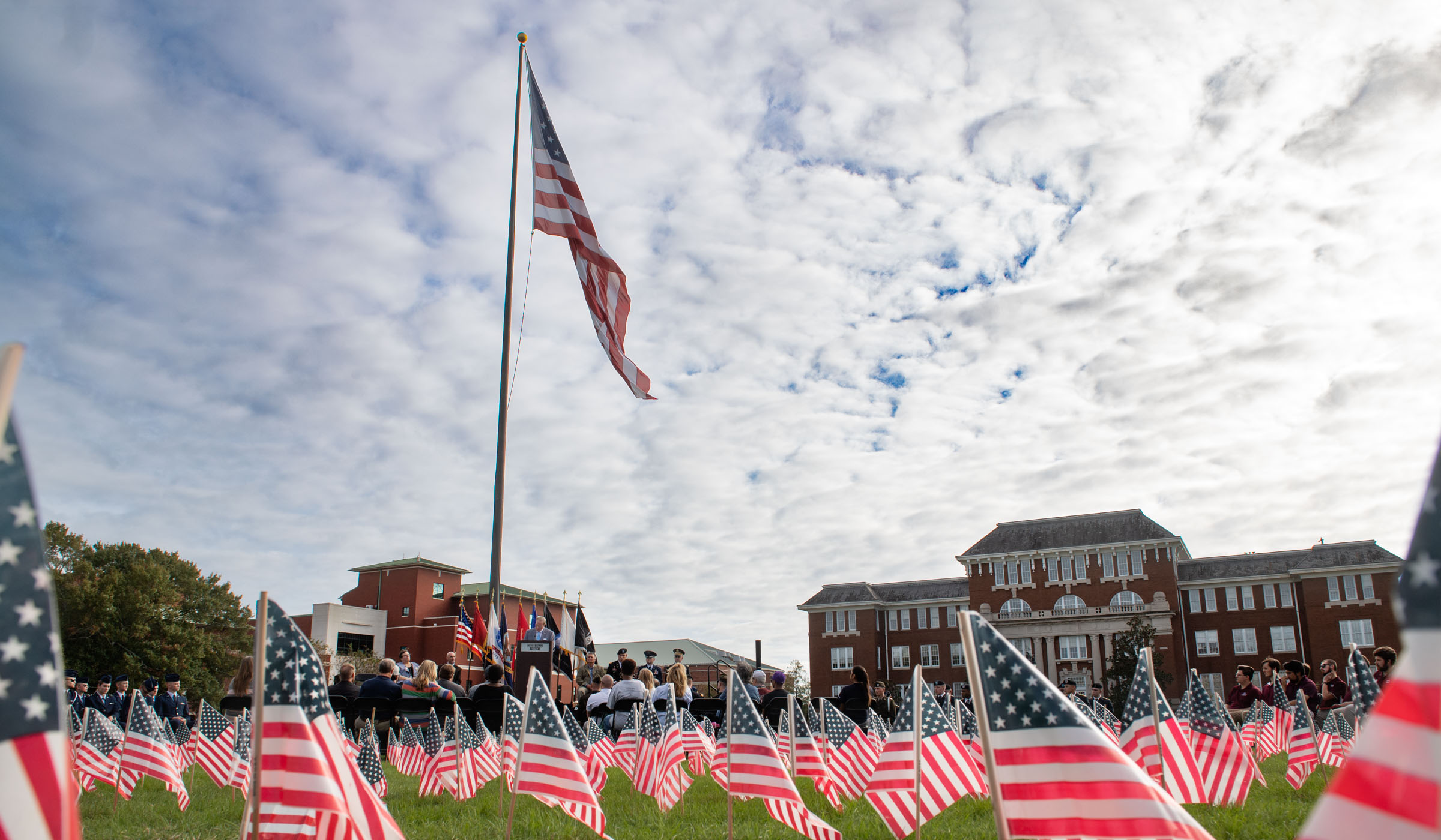 Small American flags fill the grass foreground, with the stage, Drill Field, sky and big American flag in the backfround.