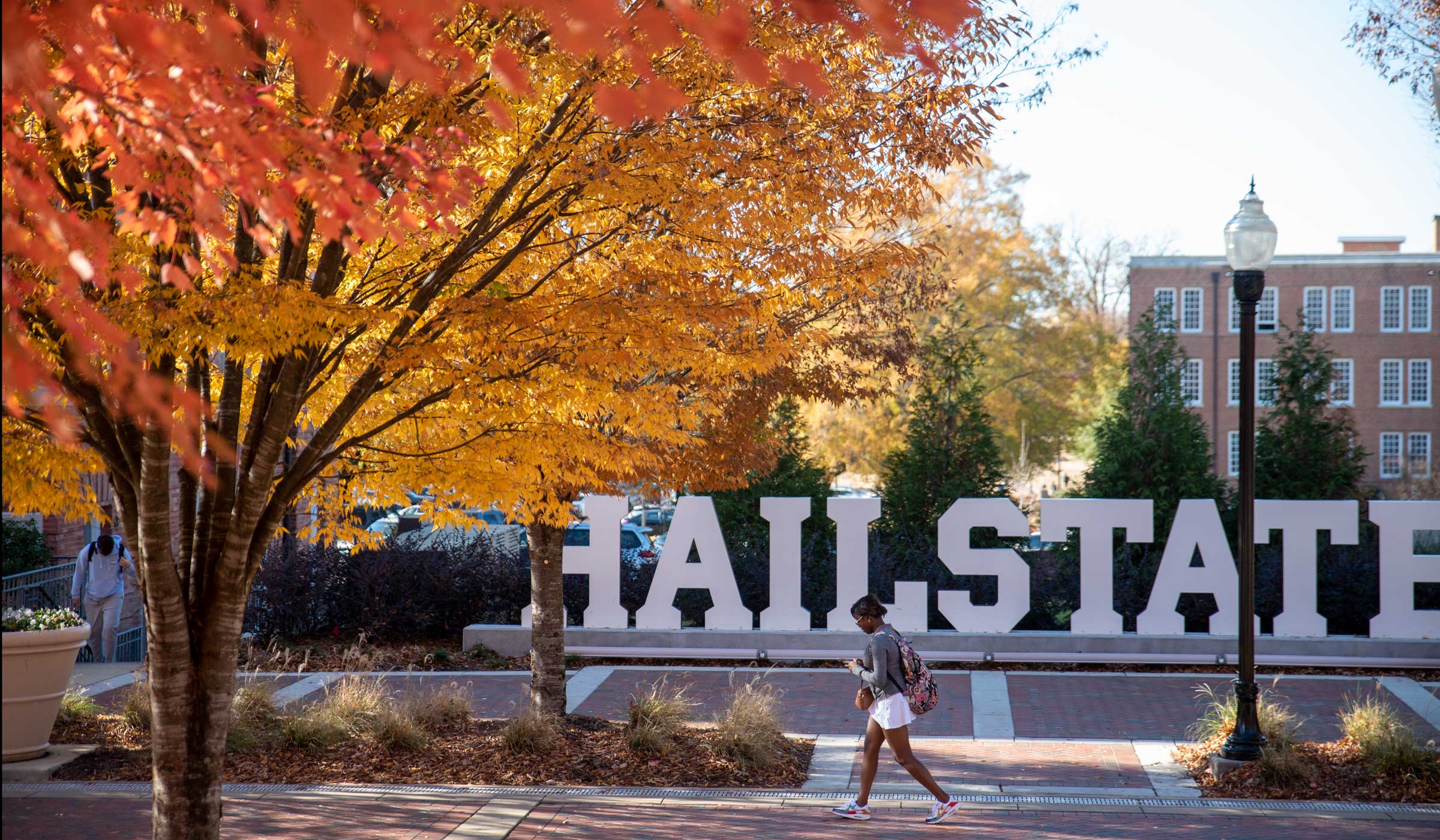 With the orange and yellow glow of fall leaves on the left, a student walks past the Hail State sign on Old Main Plaza.
