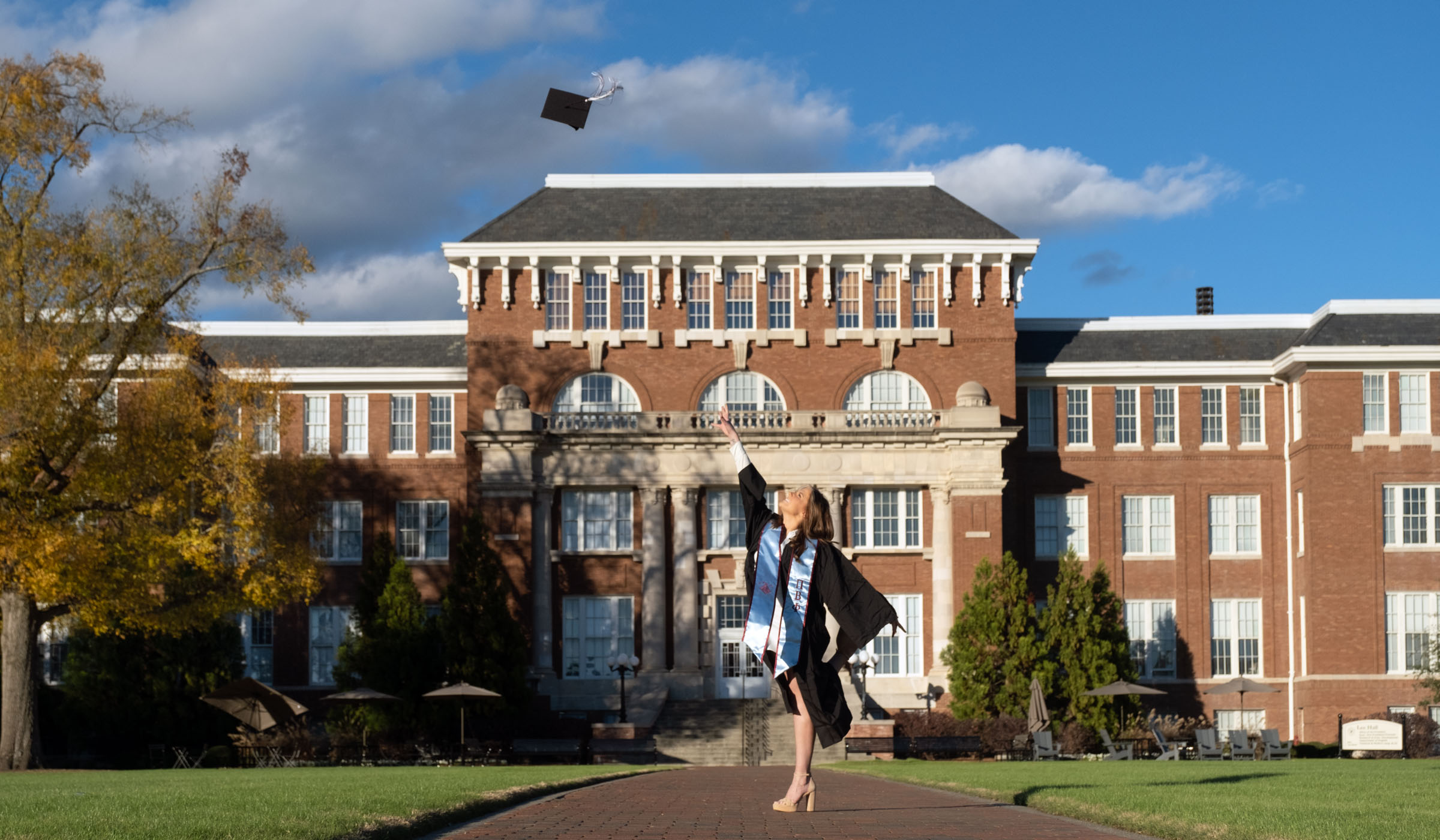 Graduate tosses cap into the air on a Drill Field sidewalk with Lee Hall in the background.